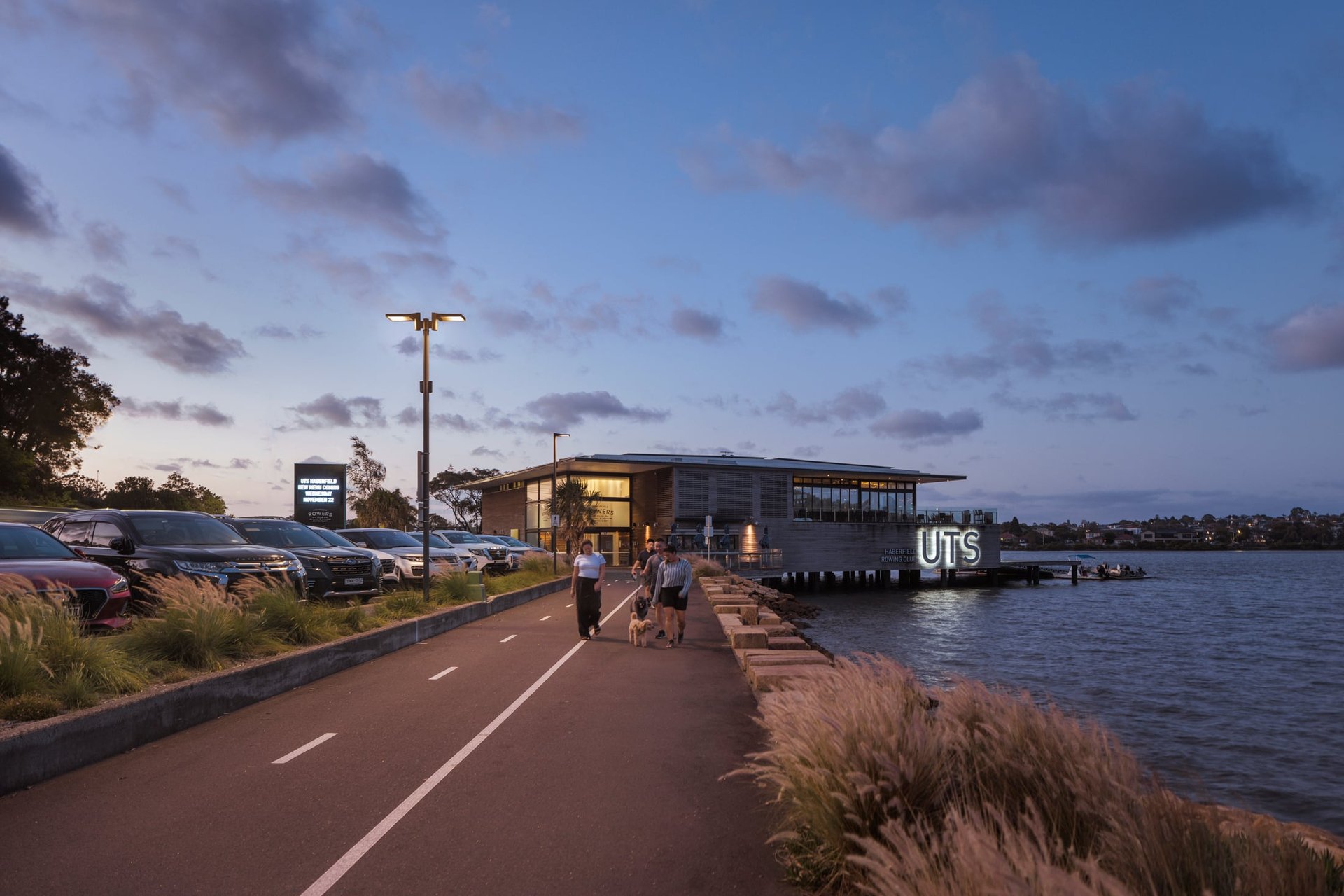 People walking a dog along Bay Run at dusk, lit by a WE-EF street luminaire. Water is on the right, parking on the left, with a city skyline in the background. The lighting upgrade improves safety and atmosphere.