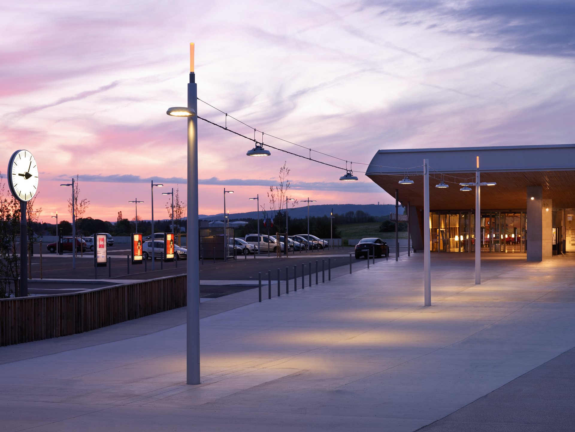 Sunset view of Belfort-Montbéliard TGV station with illuminated WE-EF RFL LED pole-mounted and catenary luminaires, station clock and warmly lit entrance building