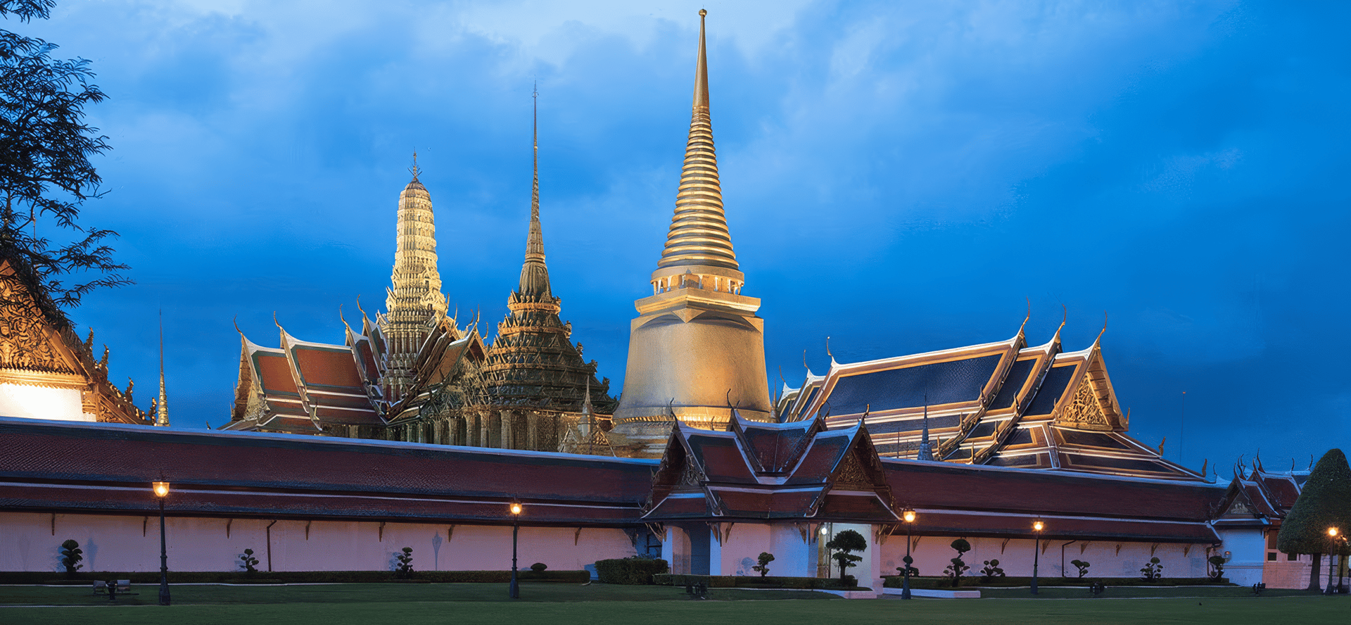 Wat Phra Kaew temple complex in Bangkok with golden stupa and spires illuminated at dusk behind the palace wall