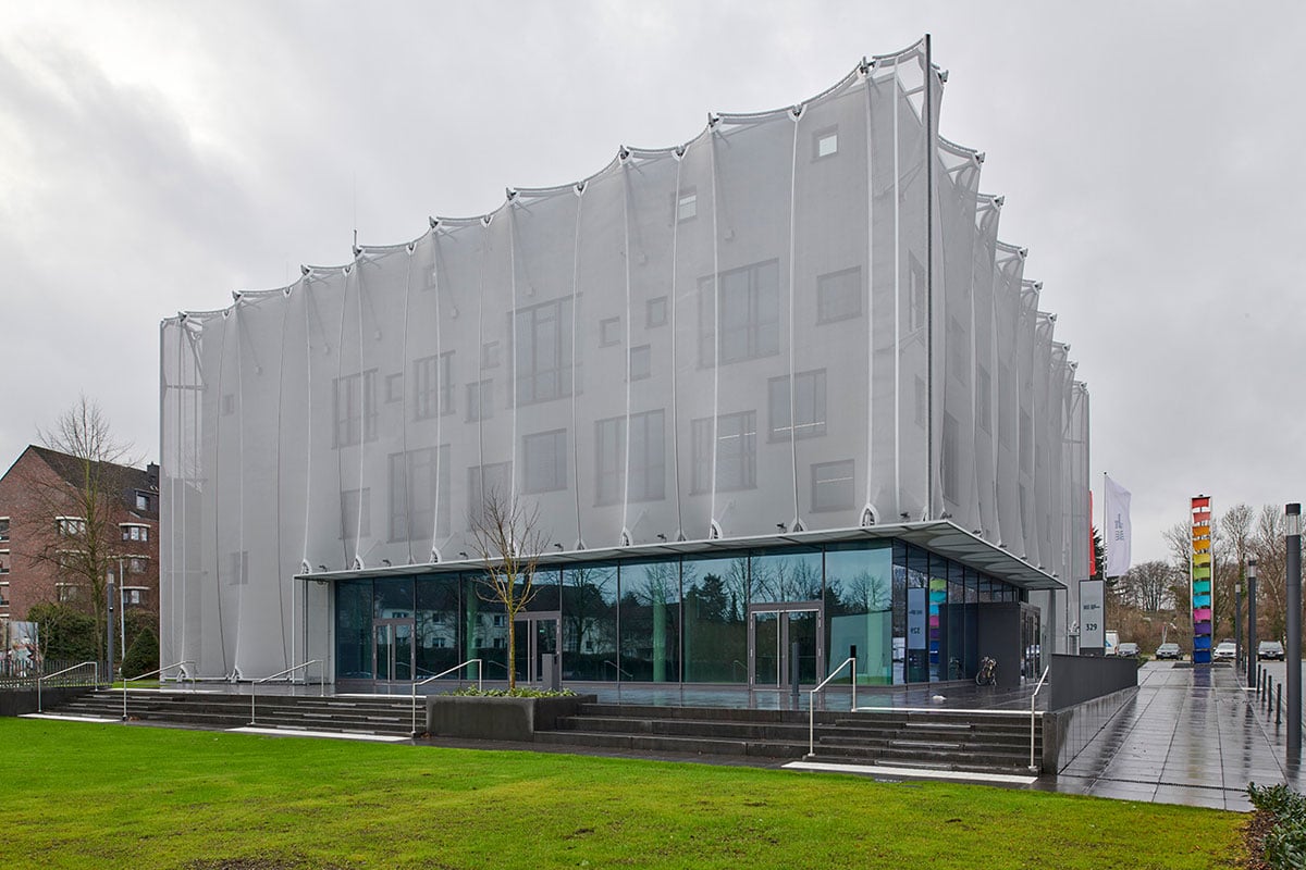 Textile Academy NRW in daylight showing shimmering metallic textile sheath appearing almost opaque despite approximately 48-percent permeability, membrane composed of individual fabric lengths creating irregular wave pattern contrasting with clear geometry of cubic structure housing central atrium, teaching and administration rooms and auditorium, window areas hidden behind membrane designed by FormTL engineers for structural and lightweight construction Radolfzell, strong symbolism implementing Mönchengladbach's long textile industry tradition through innovative architectural sheathing