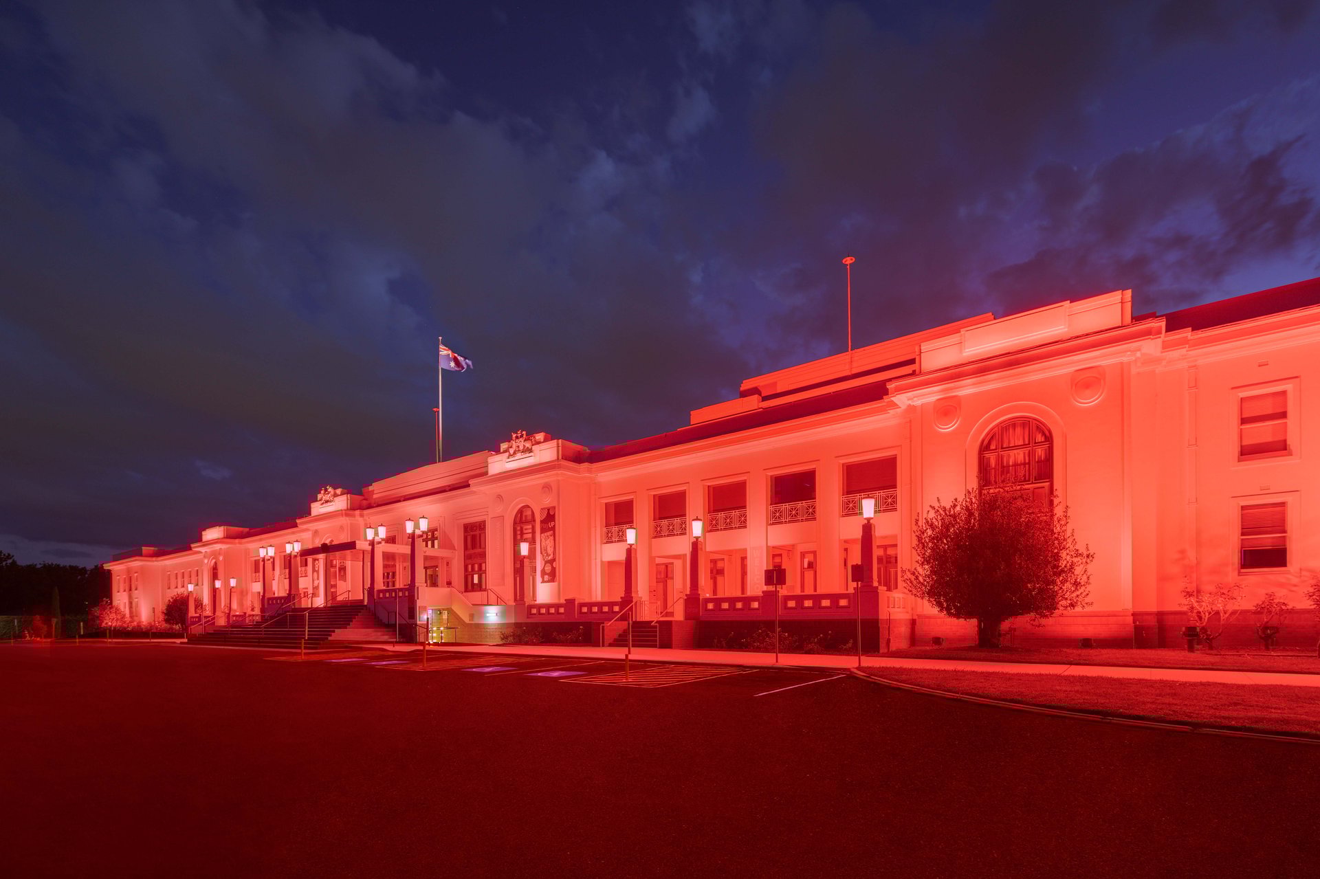 Old Parliament House front facade bathed in saturated coral-red light from WE-EF FLC200-CC and VLR100-CC linear wallwasher luminaires demonstrating superior color rendering on white exterior walls, wide-beam medium and narrow-beam optical distributions providing uniform facade coverage without visible imperfections, DMX-controlled system enabling dynamic color-changing scenarios for Museum of Australian Democracy building that housed Federal Parliament 1927-1988, Australian flag visible flying from rooftop