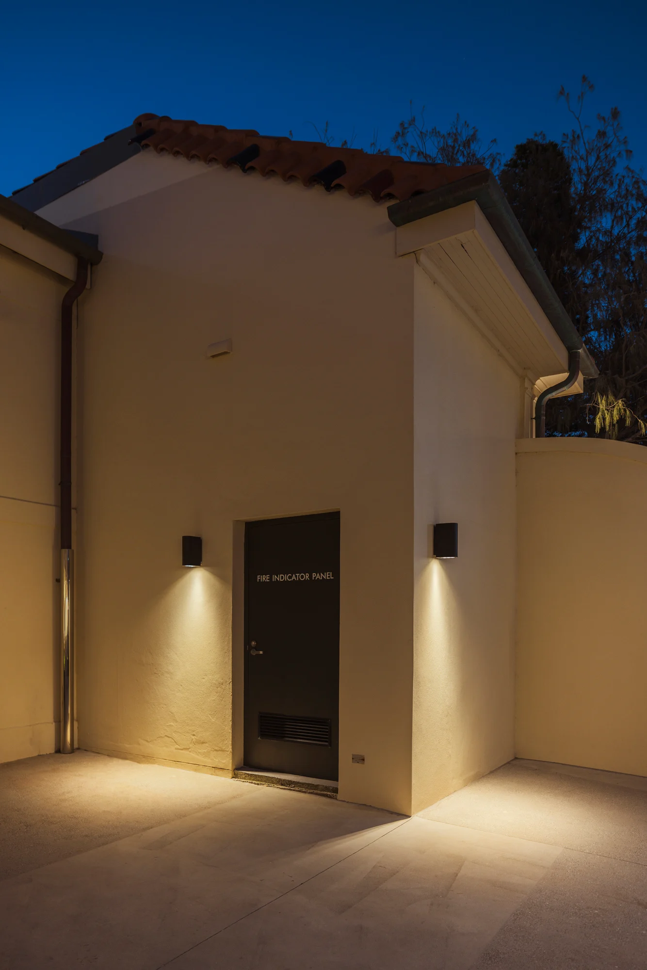 Detail of Bondi Pavilion exterior showing WE-EF wall-mounted luminaires flanking Fire Indicator Panel door, warm LED downlighting creating safe glare-free illumination on white rendered walls and terracotta roof, demonstrating heritage-sensitive lighting placement designed to accentuate architectural features while meeting strict conservation requirements for iconic beachfront cultural institution