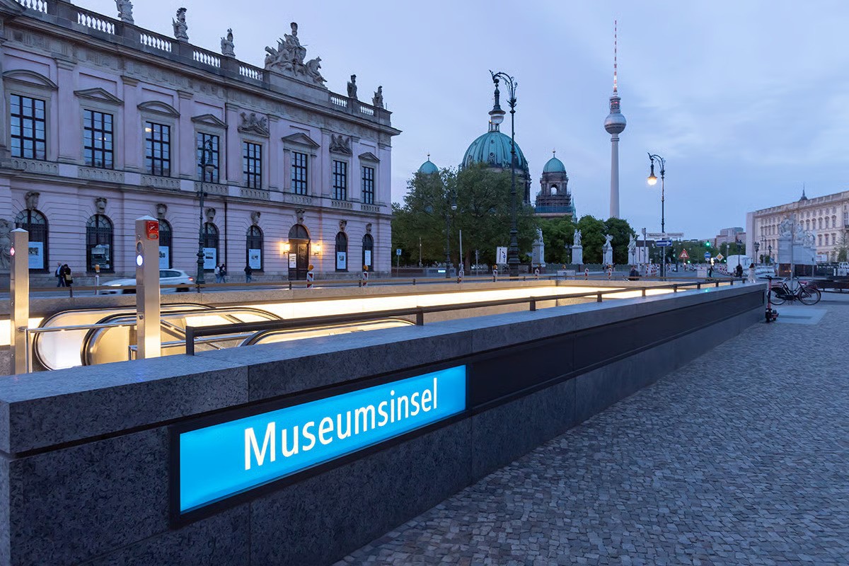 Museumsinsel U-Bahn station entrance at twilight showing contemporary open granite parapet design without traditional light portal, WE-EF VLR100 linear surface-mounted wall luminaires integrated into transparent covered wall boxes providing asymmetric wall wash downlighting into entrance, completing U5 line extension connecting Brandenburger Tor to Alexanderplatz through cultural heart of Berlin, designed by Max Dudler Architects, completed 2020 after 22-year construction period