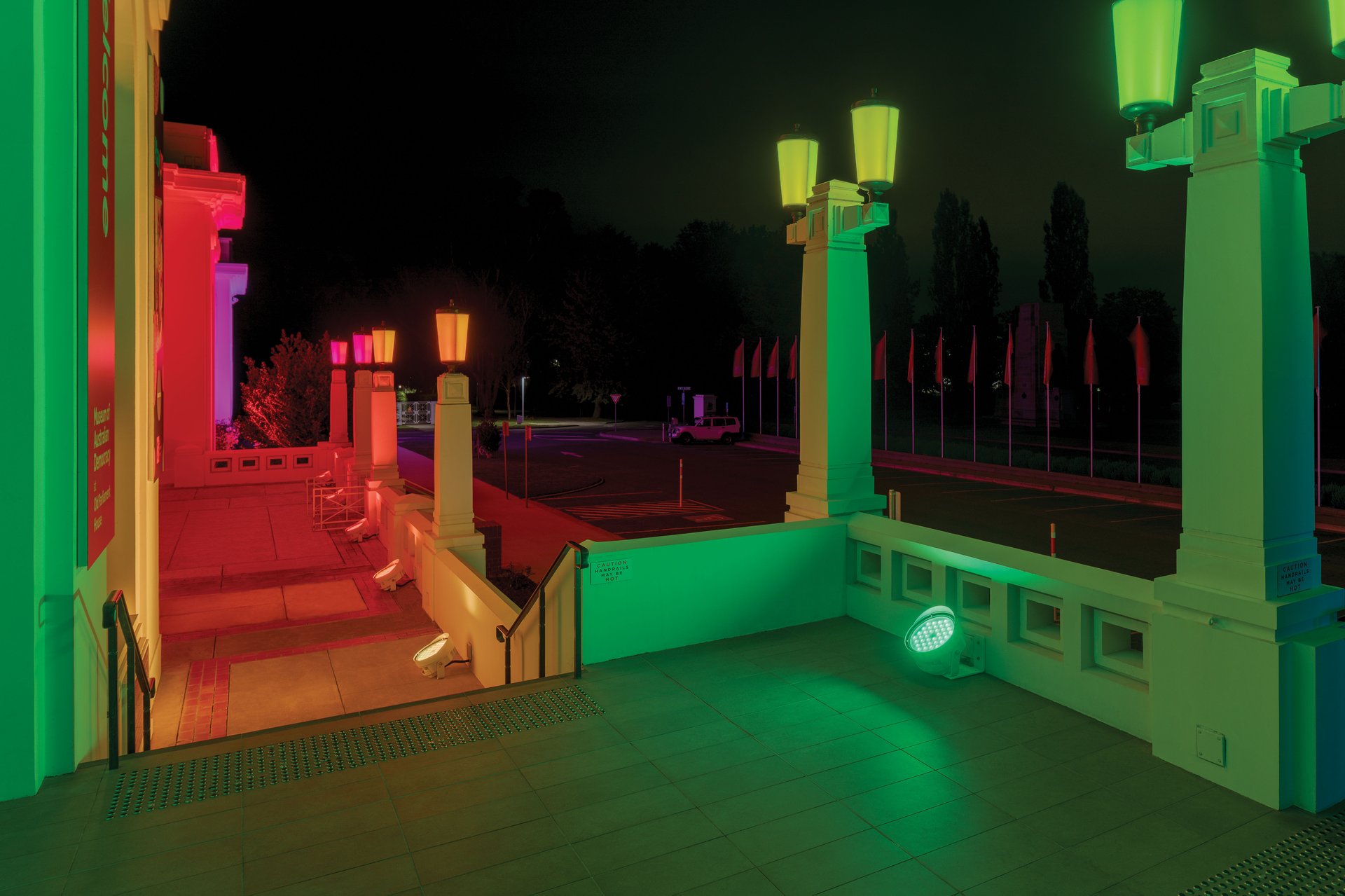 Old Parliament House rooftop terrace showing dramatic multi-color dynamic lighting effects with green, red, yellow, and purple hues from WE-EF FLC200-CC RGBW and RGBA color-changing projectors with DMX interface allowing color-changing effects to run across building, demonstrating stage lighting-based technology that paints entire heritage building in light, overcoming heritage restrictions requiring custom thin DMX cable threaded through existing small copper conduits, designed by WSP with Integral Lighting managing installation