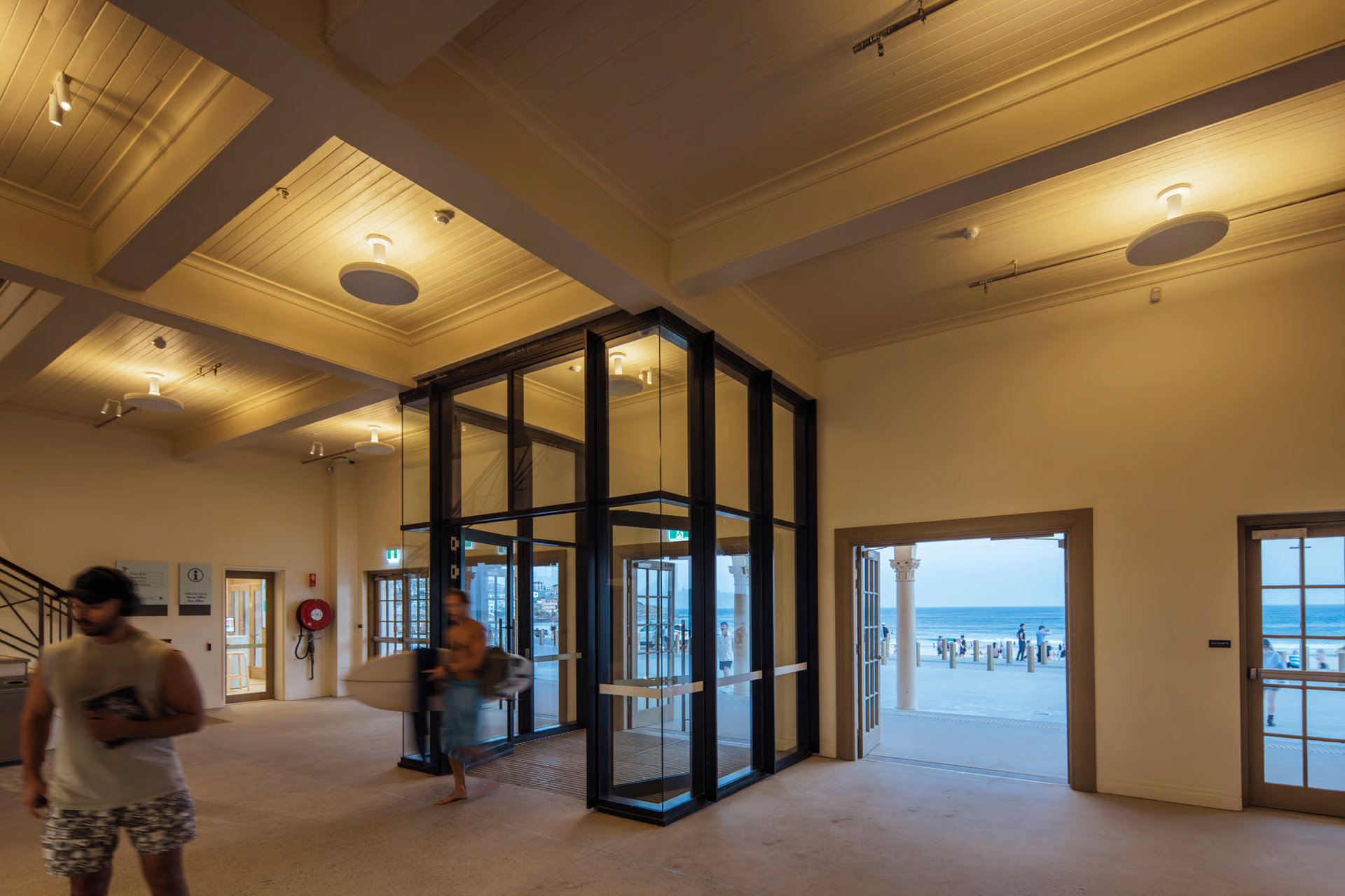 Bondi Pavilion interior lobby featuring innovative upside-down mounted WE-EF RMT320 LED bollards on coffered ceiling creating soft glare-free ambient lighting that highlights detailed timber paneling, motion-blurred visitors moving through space with black-framed glass doors opening to beachfront, demonstrating creative luminaire application in heritage building restoration designed by Electrolight