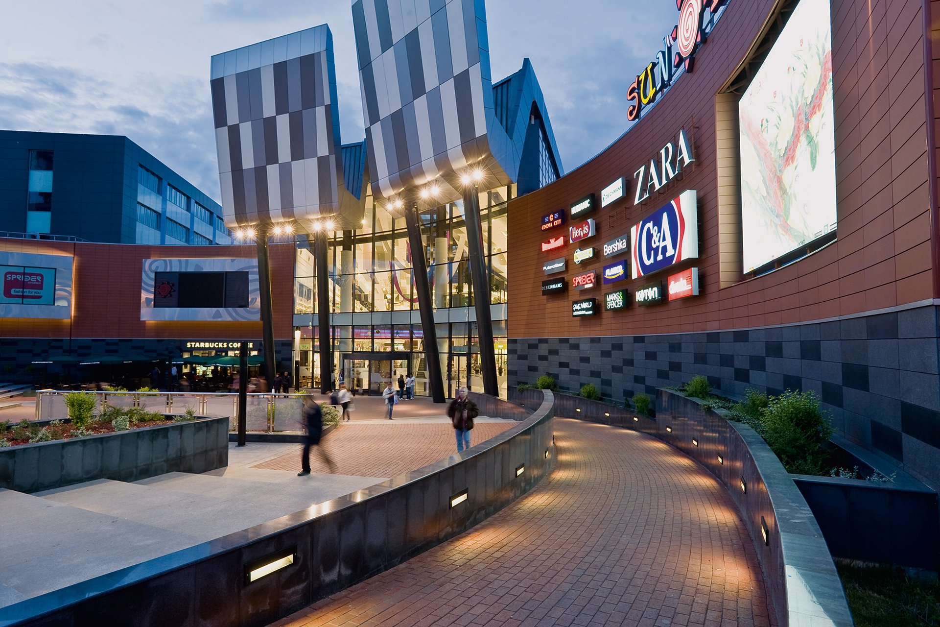 Sun Plaza shopping mall Bucharest at dusk – WE-EF RAIL66 track system with FLB141 spotlights illuminating the curved canopy of the main entrance portal, city skyline visible in the background