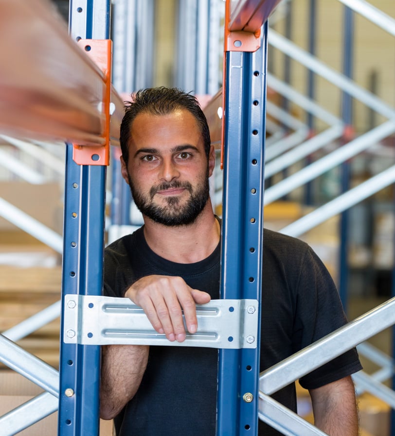 WE-EF employee in the warehouse, leaning on a blue and orange pallet rack and looking into the camera