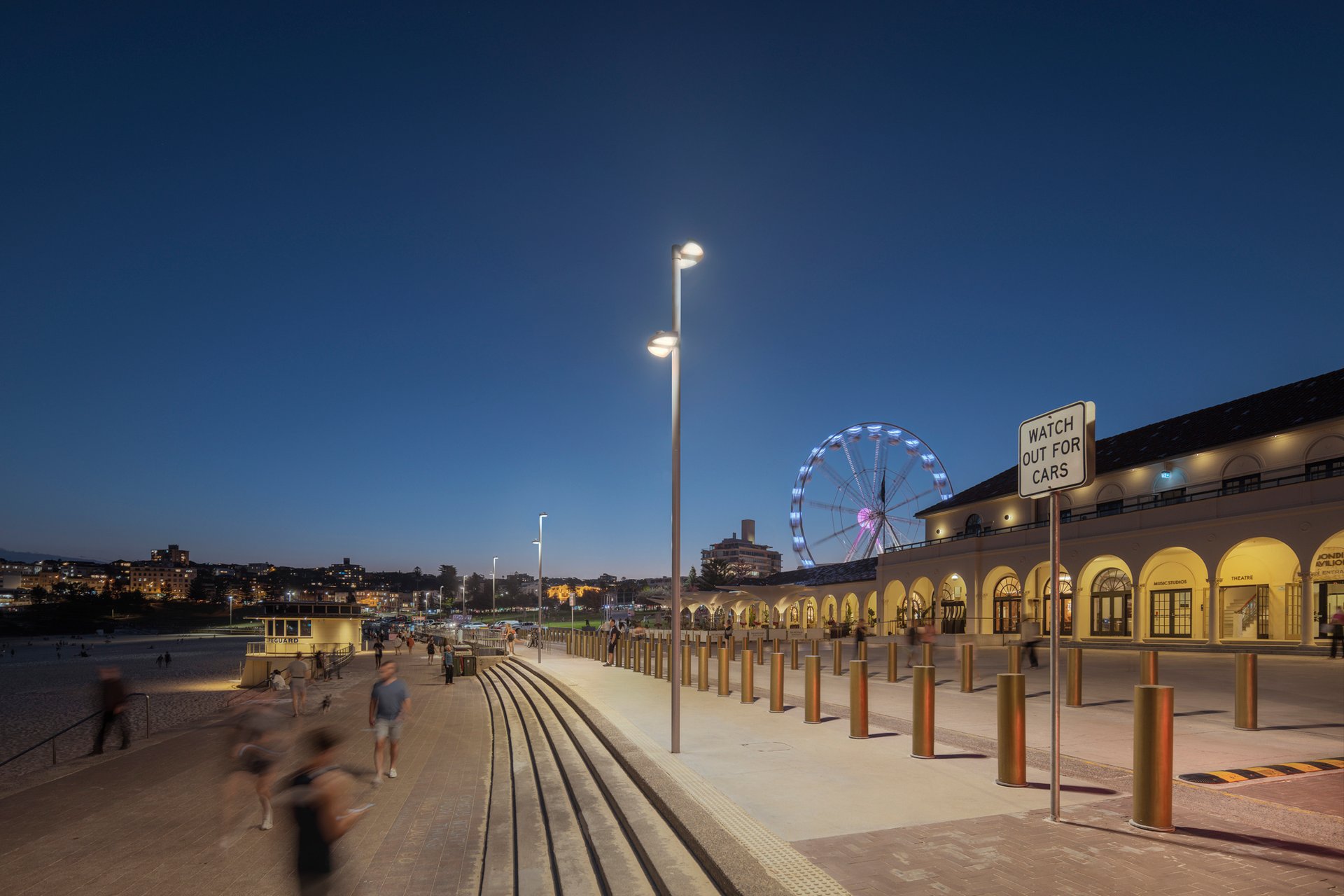 Complete view of reimagined Bondi Beach promenade showing heritage Pavilion building with multiple arched openings warmly illuminated by WE-EF luminaires, RMT320 and RMC320 bollards in bronze finish directing light strategically along pathways, VLS400 pole lights providing area illumination, illuminated Ferris wheel, motion-blurred beachgoers, demonstrating Electrolight's comprehensive lighting design ensuring pedestrian and cyclist safety while enhancing iconic Australian cultural landmark