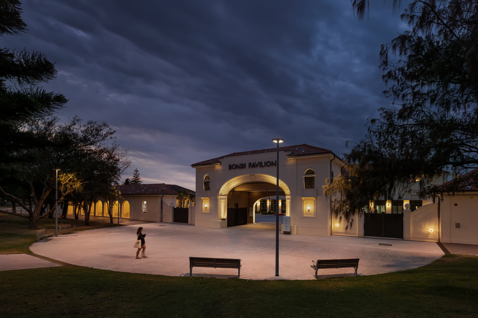 Bondi Pavilion parkland entrance at dusk showing heritage Mediterranean Revival architecture with central arched gateway, comprehensive WE-EF lighting scheme including wall luminaires creating enchanting warm 3000K ambiance throughout building and grounds, motion-blurred pedestrian on curved pathway, benches and landscaping, completed 2021 following 10+ years of stakeholder consultation and planning by Waverley Council