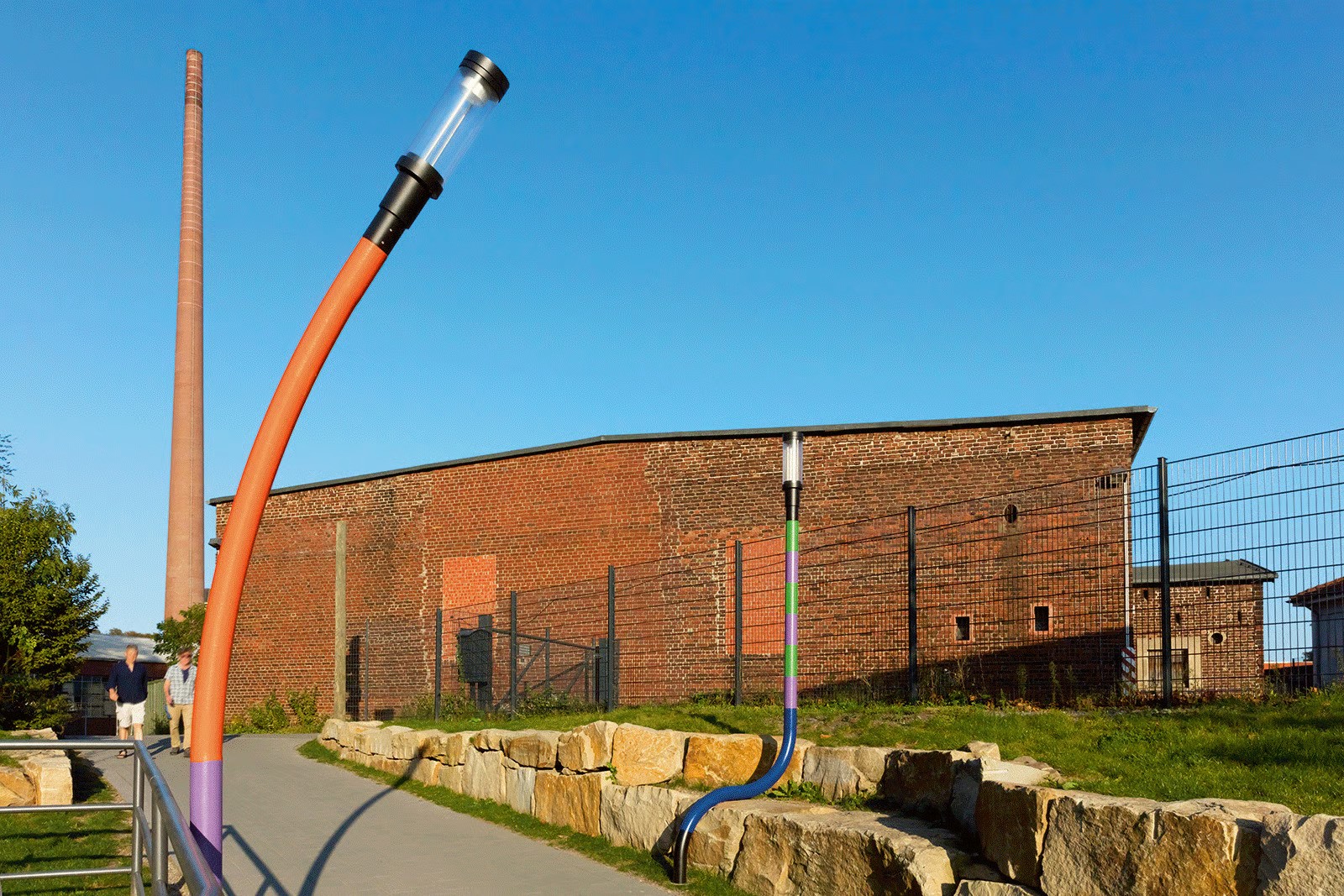 Bochum railway museum tunnel entrance showing dramatically bent orange WE-EF ZFT440 LED street luminaire in playful human-like posture designed by architect and lighting designer Peter Brdenk, illuminating both surrounding area and speech bubbles on brick wall containing information about surroundings, historic railway depot brick buildings visible behind, demonstrating quirky concept transforming formerly scary underpass into cheerful original thoroughfare giving users feeling of safety while preserving tunnel's mysterious adventurous character