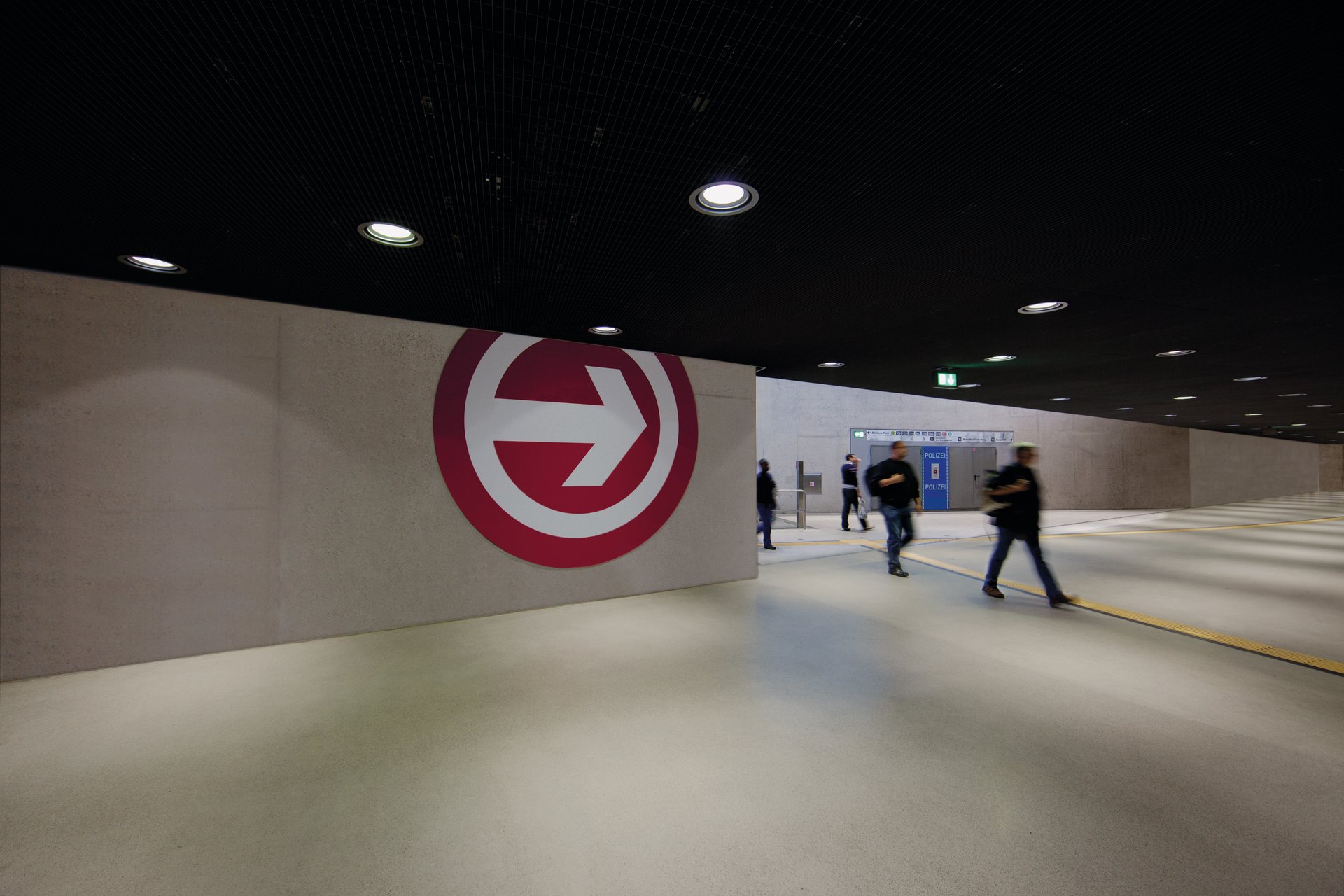 Underground concourse at Breslauer Platz station in Cologne with KVB logo on the concrete wall, WE-EF recessed ceiling luminaires and passengers in motion