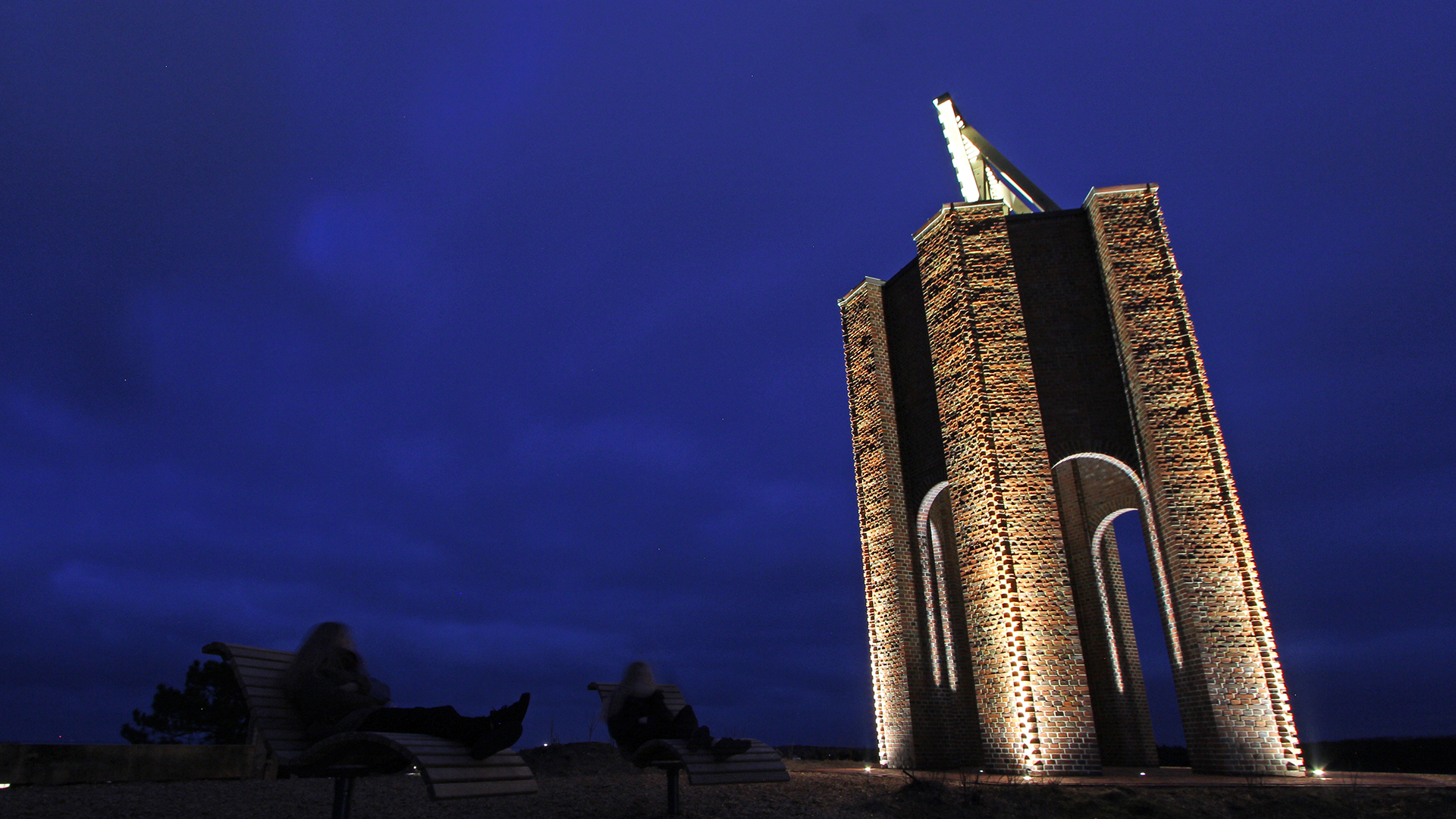 Cape Norderney historic beacon illuminated at night showing 12-meter-high hexagonal brick building erected 1871 on 15-meter beacon dune, six wall columns dramatically lit by WE-EF ETC120-GB inground luminaires with gimbal-mounted symmetric very-narrow-beam optics emphasizing lively brickwork surface texture, triangular wooden slat sea marker appearing to float above structure with round archways, symbol on Norderney island coat of arms since 1928 in East Frisian Wadden Sea serving as tourist landmark designed by Oliver Christen OC-Lichtplanung