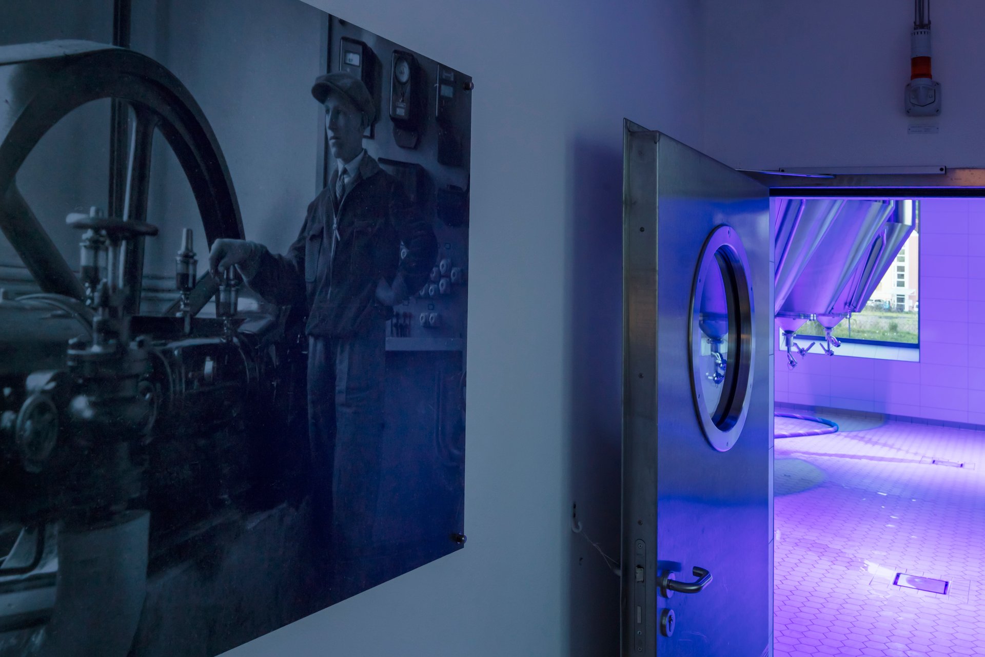 View from anteroom into Schattenhofer Brewery fermentation tank room illuminated in deep blue-violet by WE-EF FLC220-CC RGBW LED projectors, historic black-and-white photograph of traditional brewer in foreground contrasting with modern stainless-steel brewing technology and DMX-controlled color lighting designed by Bamberger engineering firm, demonstrating transformation of traditional Bavarian brewery into contemporary attraction for brewery tours