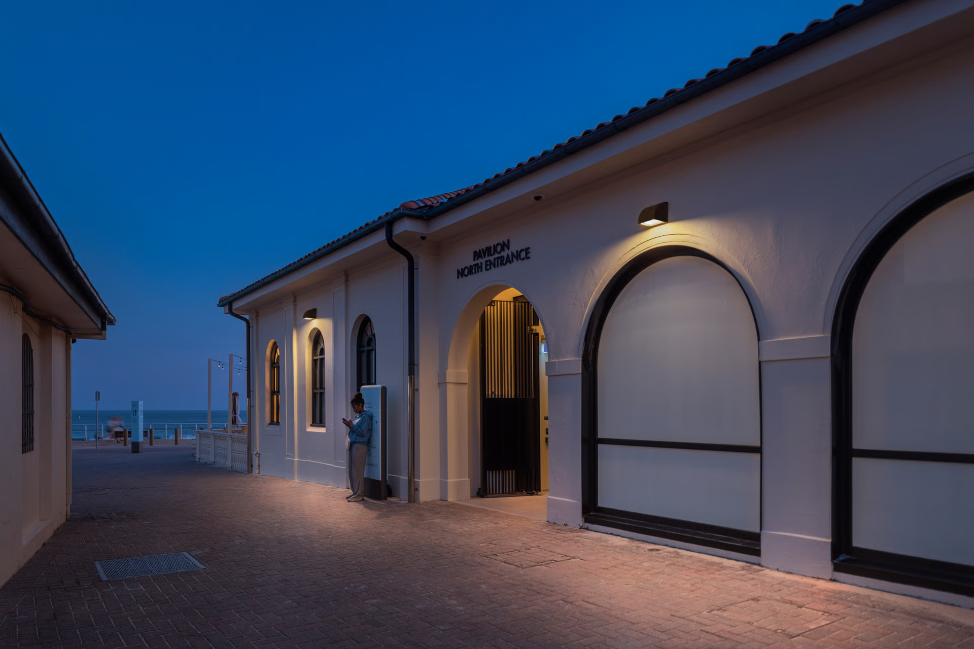 Bondi Pavilion North Entrance at twilight showing heritage-listed white stucco facade with terracotta tile roof and arched openings, WE-EF OLV300 wall luminaires with customized optics illuminating arch windows, warm 3000K LED lighting highlighting architectural features while avoiding upward light spill, designed by Electrolight and Tonkin Zulaikha Greer Architects as part of decade-long heritage restoration project