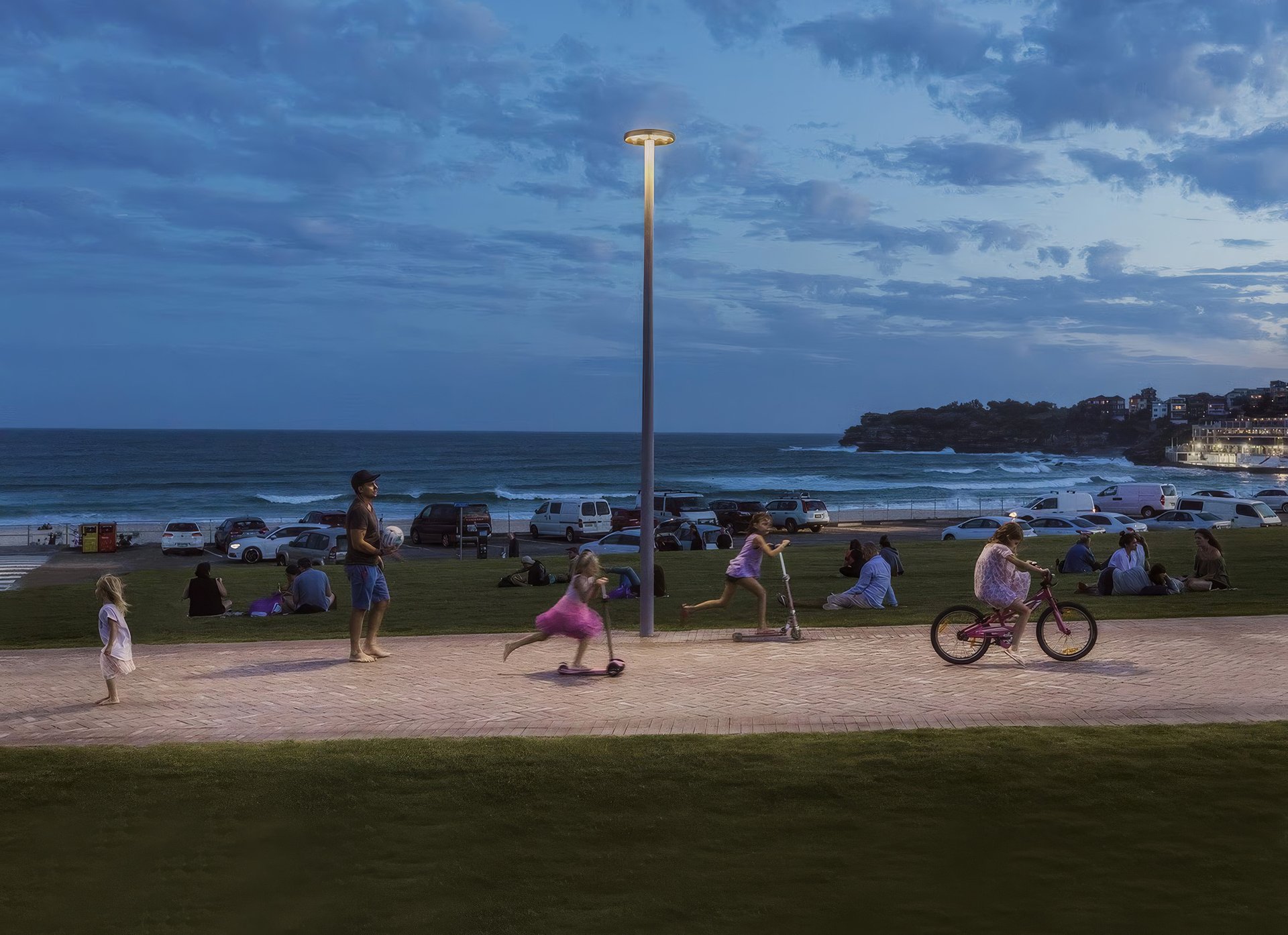 WE-EF light column illuminating a seaside path at Bondi Beach at dusk, with children riding scooters and bikes