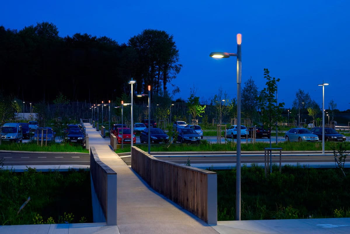Nachtaufnahme des beleuchteten Fußwegs am Parkplatz des TGV-Bahnhofs Belfort-Montbéliard mit WE-EF RFS530-LED-Mastleuchten, geschwungener Holzbrüstung und weitläufigem Parkbereich im Hintergrund