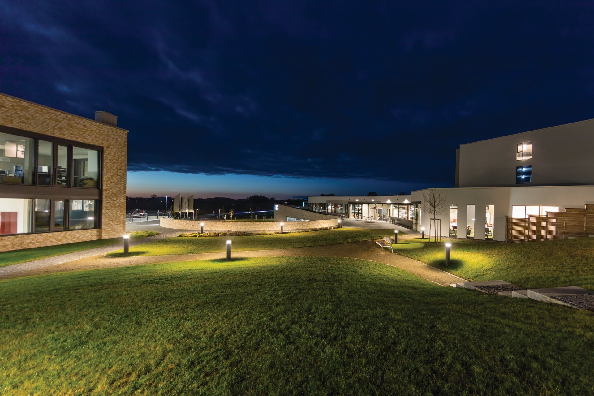 das flax Hotel illuminated at night in Allgäu Alpine foothills showing campus-style architecture by FG-Architektur with multiple buildings around central circular space, complete WE-EF lighting solution featuring KTY234 pathway bollards, VFL530 dual-arm driveway luminaires, ETV130 linear facade washers, and ETC120-GB gimbal-adjustable tree uplights, unified 3000K warm white LED system supporting bleisure travel concept combining business functionality with welcoming leisure atmosphere in sustainable economy hotel completed 2020