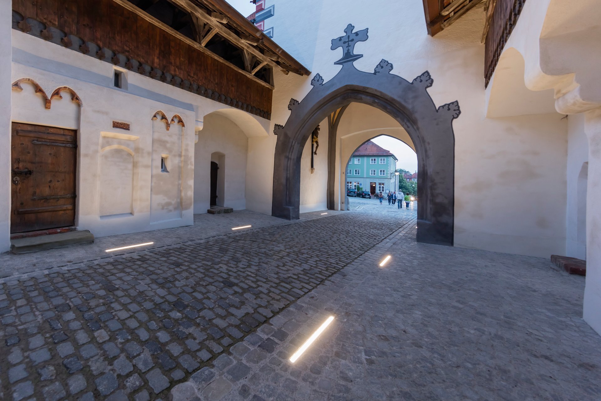 Bayertor's medieval courtyard at twilight featuring white plastered walls with Gothic arched doorways, traditional wooden balconies with terracotta roof tiles, and WE-EF ETV130 linear luminaires creating vertical wallwashing that highlights architectural details