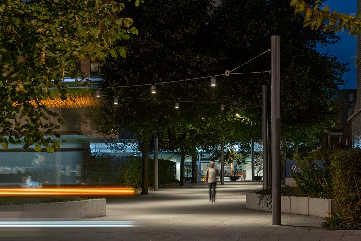 Southern House entrance at night showing full effect of custom WE-EF DAC220 catenary lighting system on specially engineered taut wire cables, symmetric wide-beam LED optics with CAD-optimized OLC technology providing superior glare control, warm 3000K LED creating inviting individually illuminated space as pedestrian connection between East Croydon station and rejuvenated office complex, completed 2018 for LCR Property
