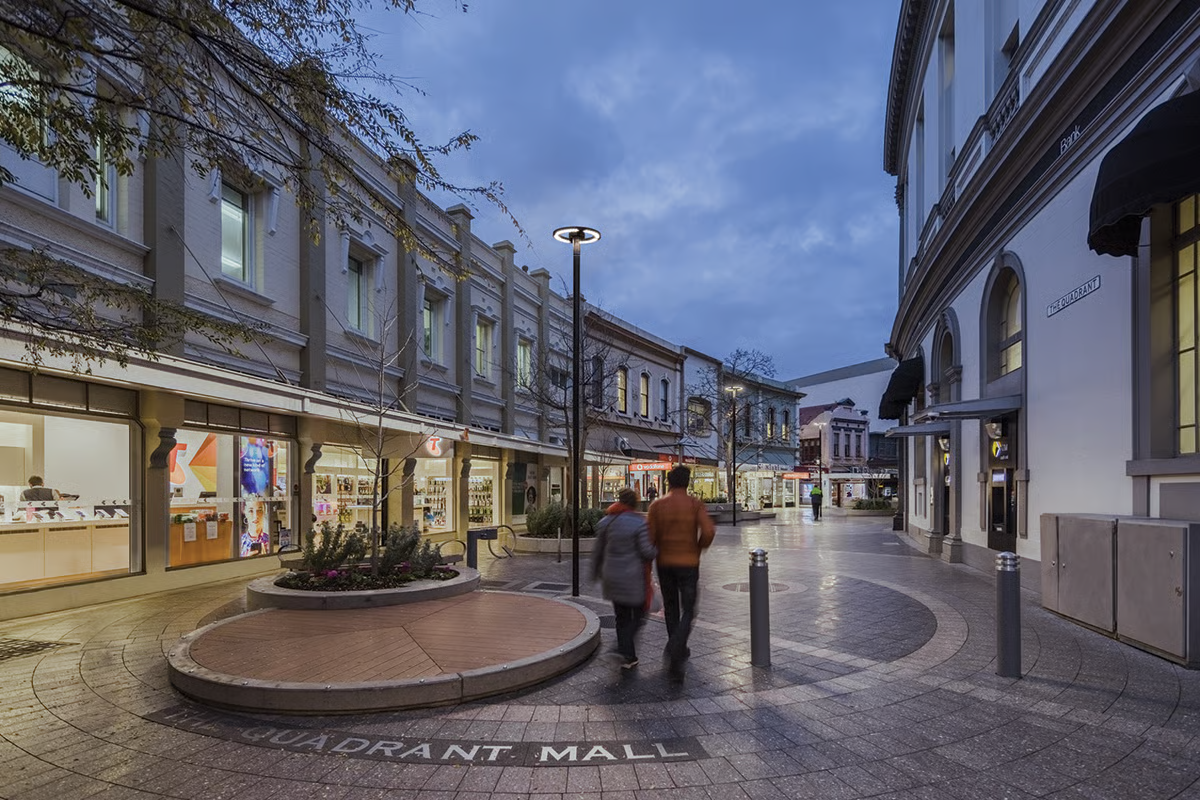 Quadrant Mall pedestrian shopping precinct in Launceston at dusk, featuring motion-blurred shoppers, WE-EF CFT540 LED pole lights with distinctive circular luminaires, curved planters with landscaping, graphic paving patterns, and heritage Victorian buildings housing retail stores along the revitalized City Heart Project streetscape