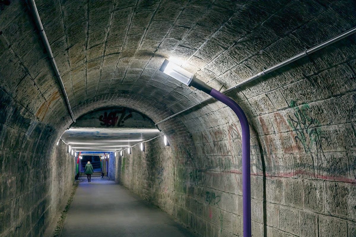 IBochum railway museum pedestrian tunnel interior showing 114-meter-long 100+ year-old underpass with historic stone walls bearing patina, arched vault transitioning to low flat ceiling sections, individually colored purple pole with WE-EF ZFT440 LED luminaire (symmetric C60 beam, 4000K) bent in quirky human-like posture, 26 WE-EF DLO239 LED wall luminaires providing pleasant illuminances, silhouetted walker demonstrating transformed safe attractive passage connecting Dahlhausen district with Ruhr river meadows and cycle pathsmage