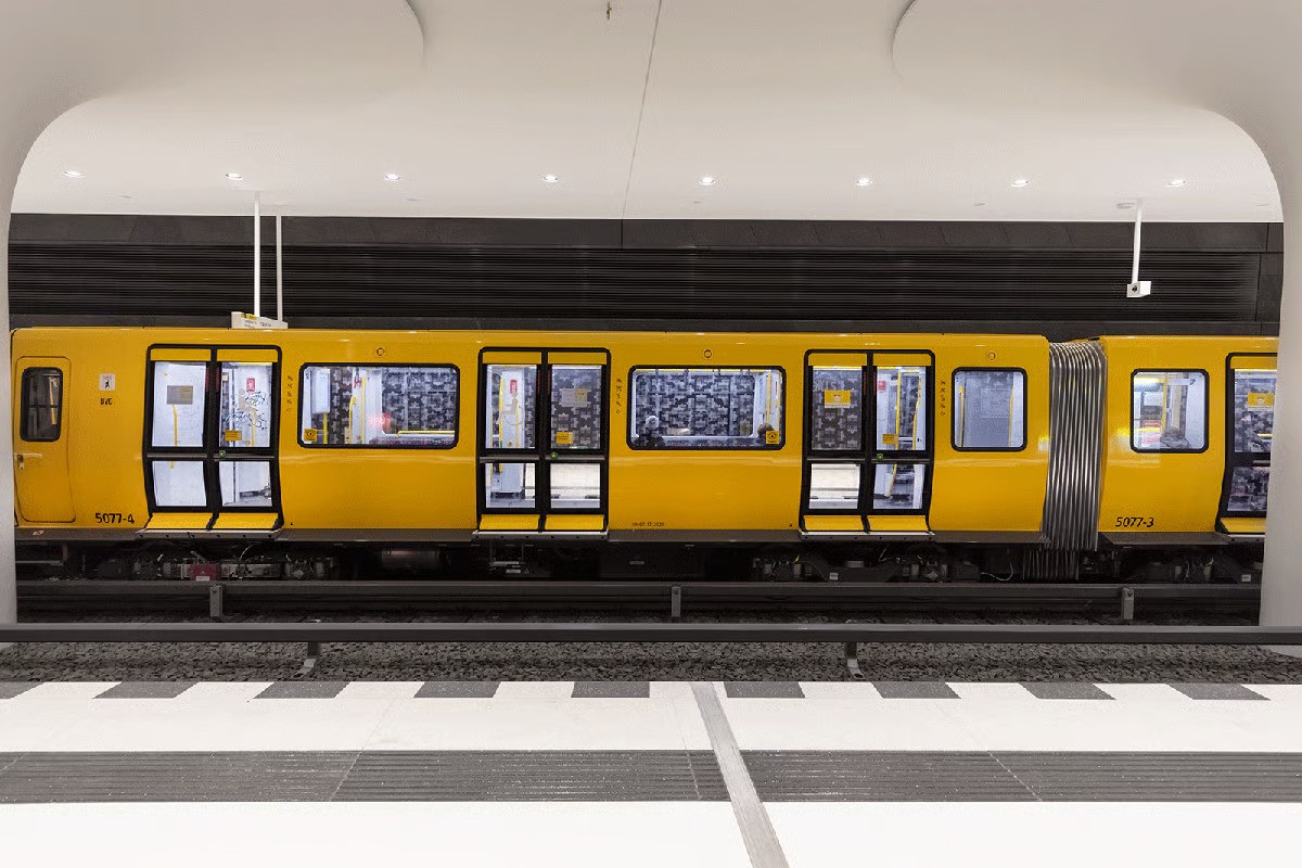 Rotes Rathaus U-Bahn station platform showing striking black and white composition with gleaming white floors and ceiling, dark polished concrete walls with subtle sparkle, yellow Berlin BVG train, 70 WE-EF DOC240 LED recessed ceiling luminaires with 24 LEDs in warm white 3000K and 48W connected load producing symmetrical medium-beam distribution, reflection from white surfaces creating pure lighting effect with high visual comfort, completed December 2020 at seat of Berlin state government