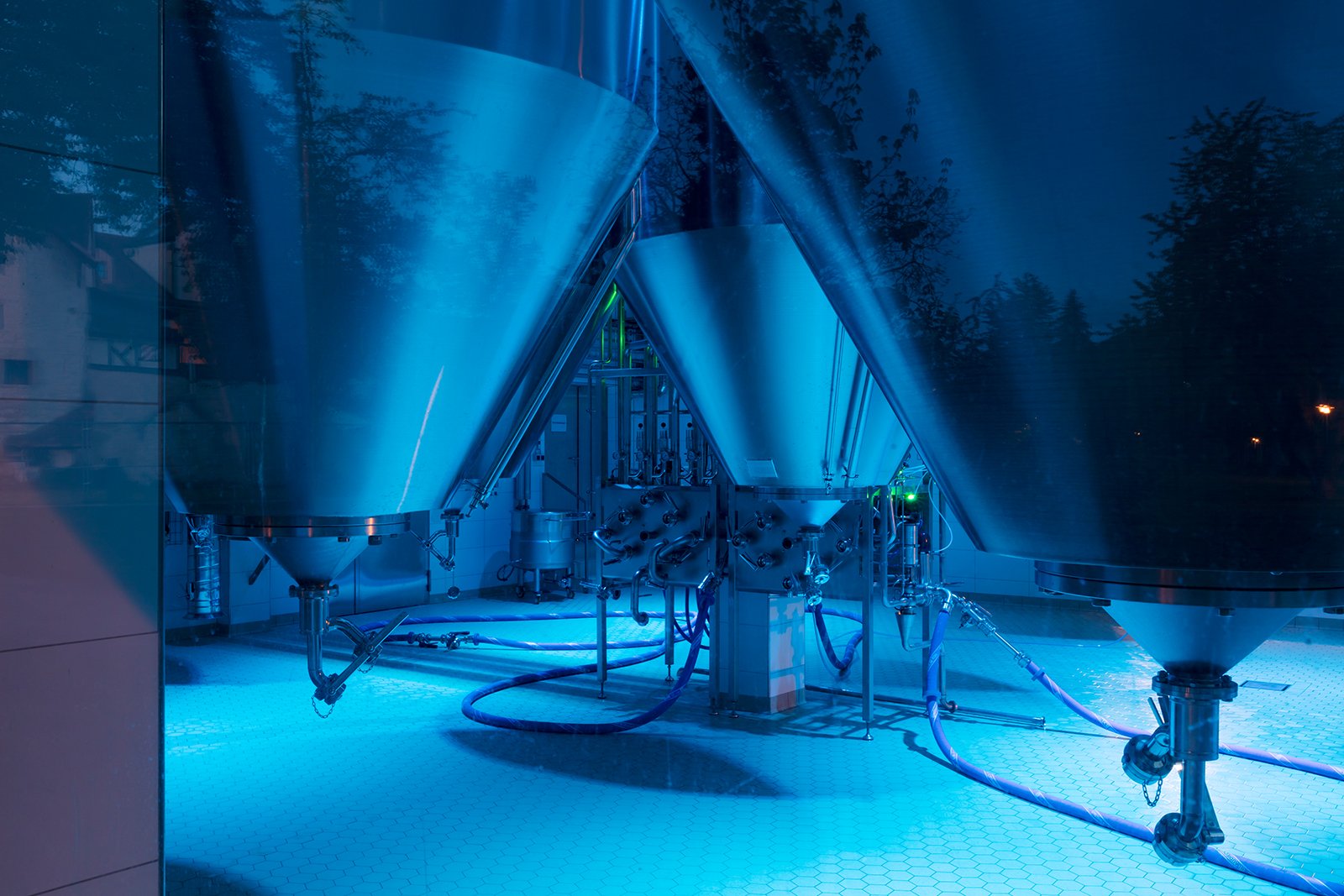 Schattenhofer Brewery fermentation tank room at night showing atmospheric cyan-blue lighting from WE-EF FLC220-CC RGBW projectors with IP66 outdoor-rated housing creating magical spectacle visible through panoramic windows, stainless-steel tanks and brewing equipment transformed into dramatic visual attraction with timer-activated automatic color progressions, demonstrating how forward-thinking 300-year-old Bavarian family brewery signals transparency, quality awareness and modernity through futuristic architectural lighting