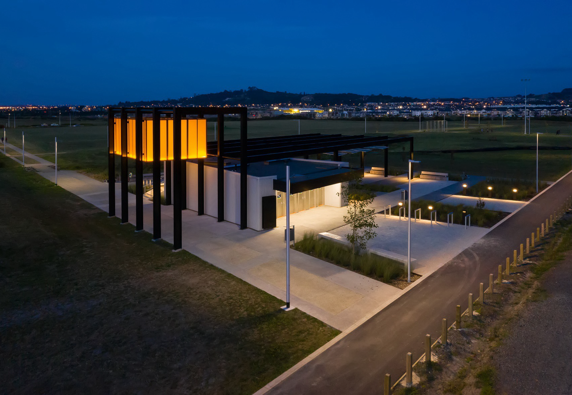 Barry Curtis Park award-winning pavilion at night in Flat Bush Auckland showing dramatic orange lantern design honoring local Chinese community while meeting flight path visibility requirements, comprehensive WE-EF lighting by MHL including FLC100 projectors creating shadow play on layered ceiling, QSI254 bollards at changing rooms, 150 STG200 recessed steplights in pre-cut walls, largest urban park developed in Auckland in past century at 25km south-east of city center integrating sports and recreation with protected wetland habitat for native birdlife and fauna, completed 2018 as first stage of multi-year masterpiece for Auckland Council