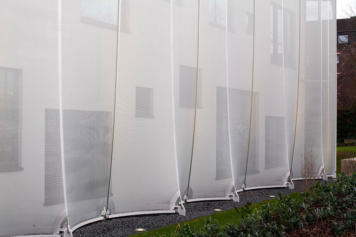 Close-up detail of Textile Academy NRW approximately 2,100-square-meter shimmering silver membrane sheath showing individual fabric lengths alternately stretched in front of and behind ropes forming irregular wave pattern, flowing form nestling around functional building structure contrasting clear three-story cube geometry, innovative textile facade reacting to changing light conditions creating distinctive night-time look when WE-EF uplighting reveals irregularly distributed windows of different sizes visible under modeled membrane, demonstrating complex design language showcasing textile industry professions taught at private vocational college