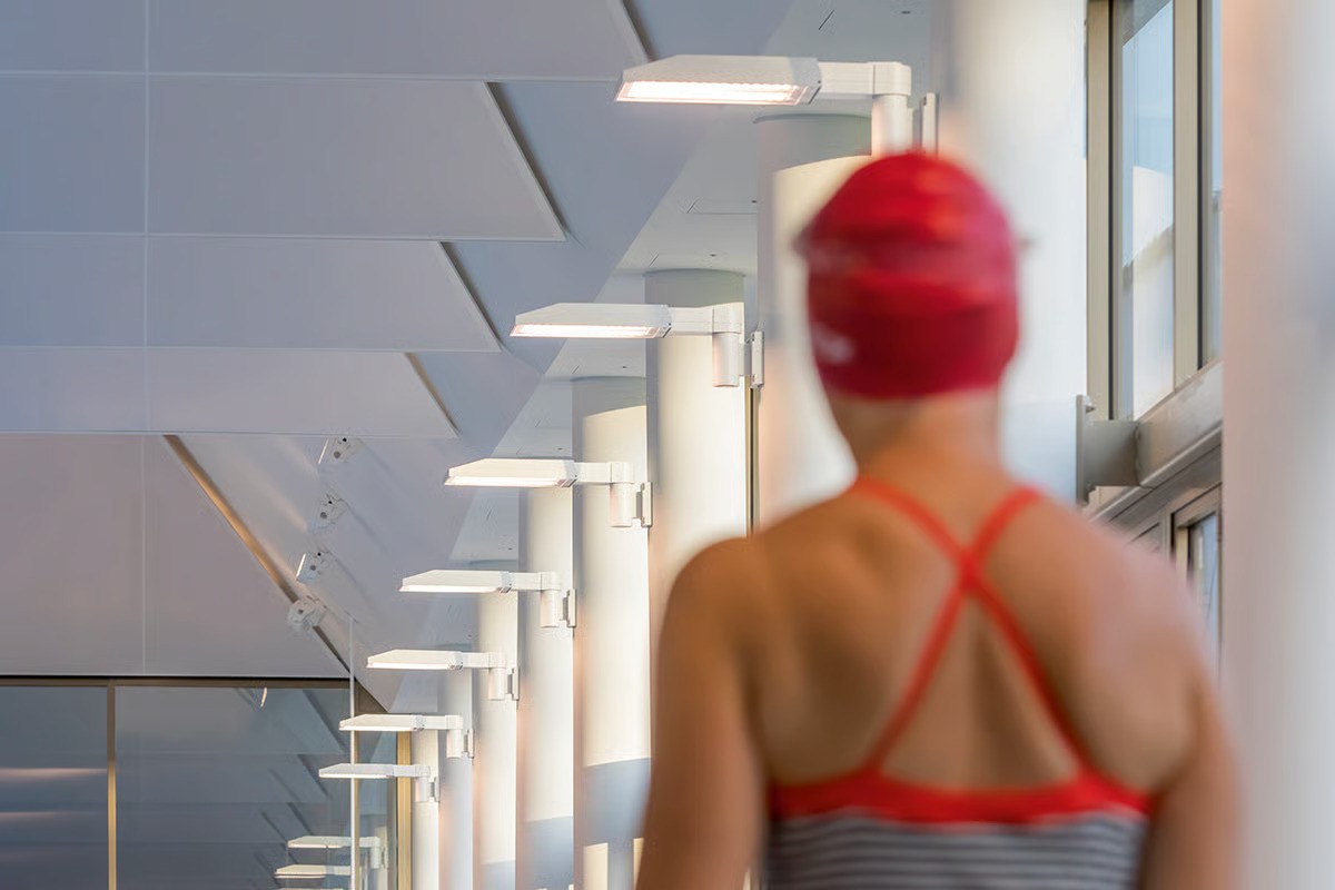 Hamburg-Ohlsdorf family swimming pool interior showing swimmer in red swimsuit and cap with WE-EF FLC230 LED projectors creating dramatic grazing light effect on folded textile acoustic ceiling panels using extremely narrow-beam optics with linear spread lens, warm 3000K color temperature, wooden truss ceiling structure concealed behind light acoustic membrane elements, DALI-controlled lighting with integrated emergency lighting monitoring modules in select luminaires
