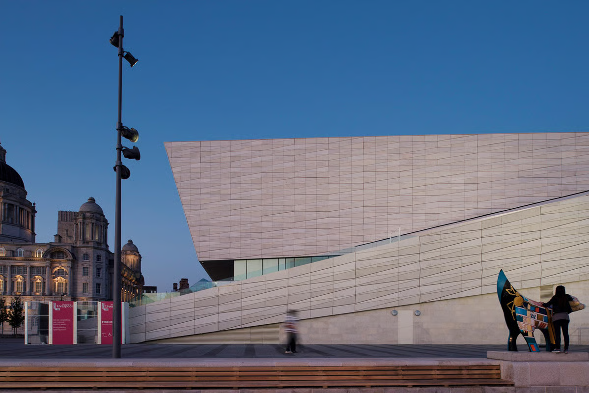 Dusk view of the National Museum Liverpool with its illuminated limestone facade, FLC projectors on a pole in the foreground, a colourful sculpture at the entrance, and the historic Pier Head buildings in the background