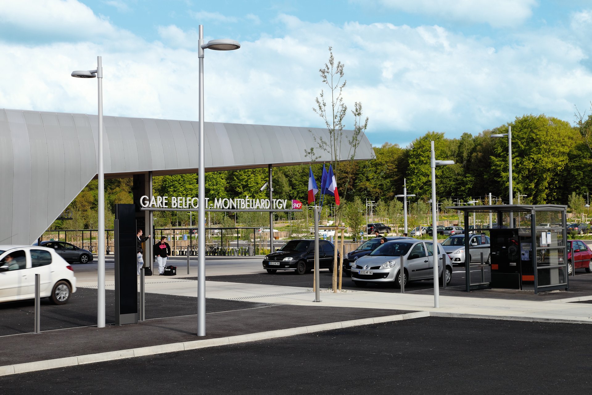 Tagesaufnahme des TGV-Bahnhofs Belfort-Montbéliard mit dem Stationsschriftzug, WE-EF Mastleuchten auf dem Vorplatz, Pkw-Stellplätzen und französischen Flaggen