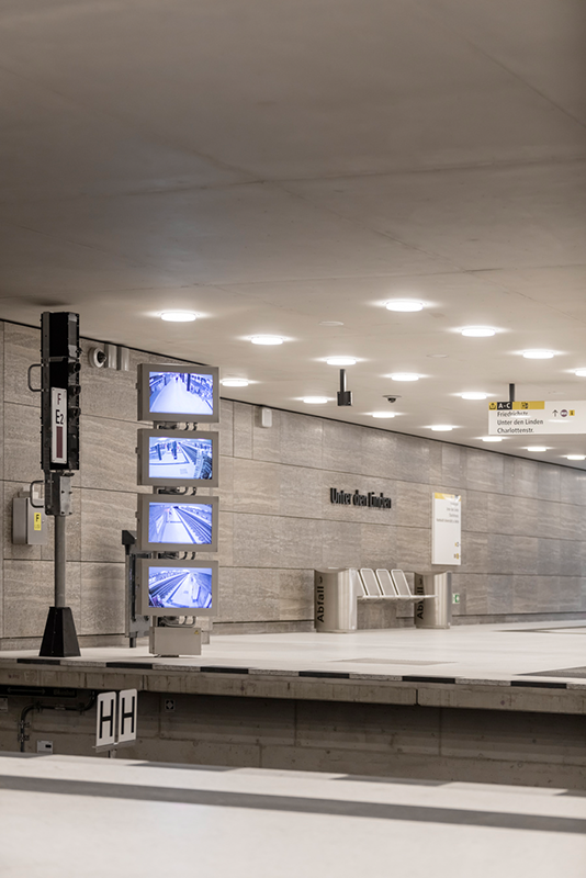 Unter den Linden U-Bahn platform level showing shell limestone wall cladding with digital information displays, 410 WE-EF LED recessed ceiling luminaires providing uniform soft 3000K warm white illumination meeting strict transport infrastructure standards, project-specific DOC240 luminaires with modified mounting elements optimized for installation cavities created years earlier during cut-and-cover construction, completed 2020 for BVG Berliner Verkehrsbetriebe completing prestigious U5 line extension