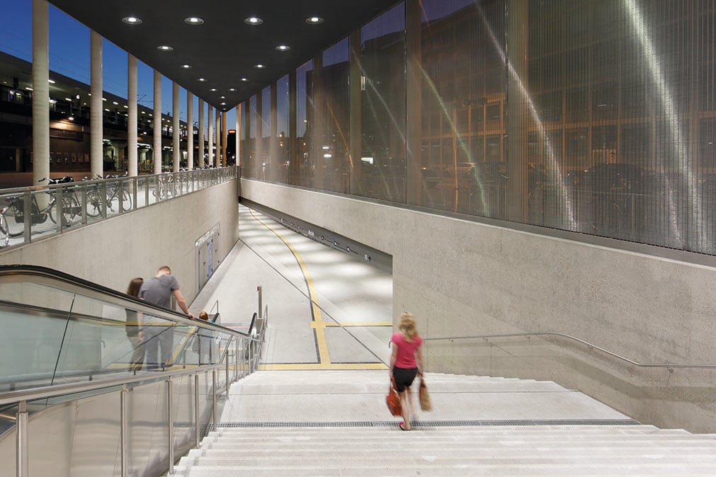 Stairway and escalator entrance at Breslauer Platz underground station in Cologne at night, with WE-EF recessed ceiling luminaires, a structured glass facade and passengers in motion