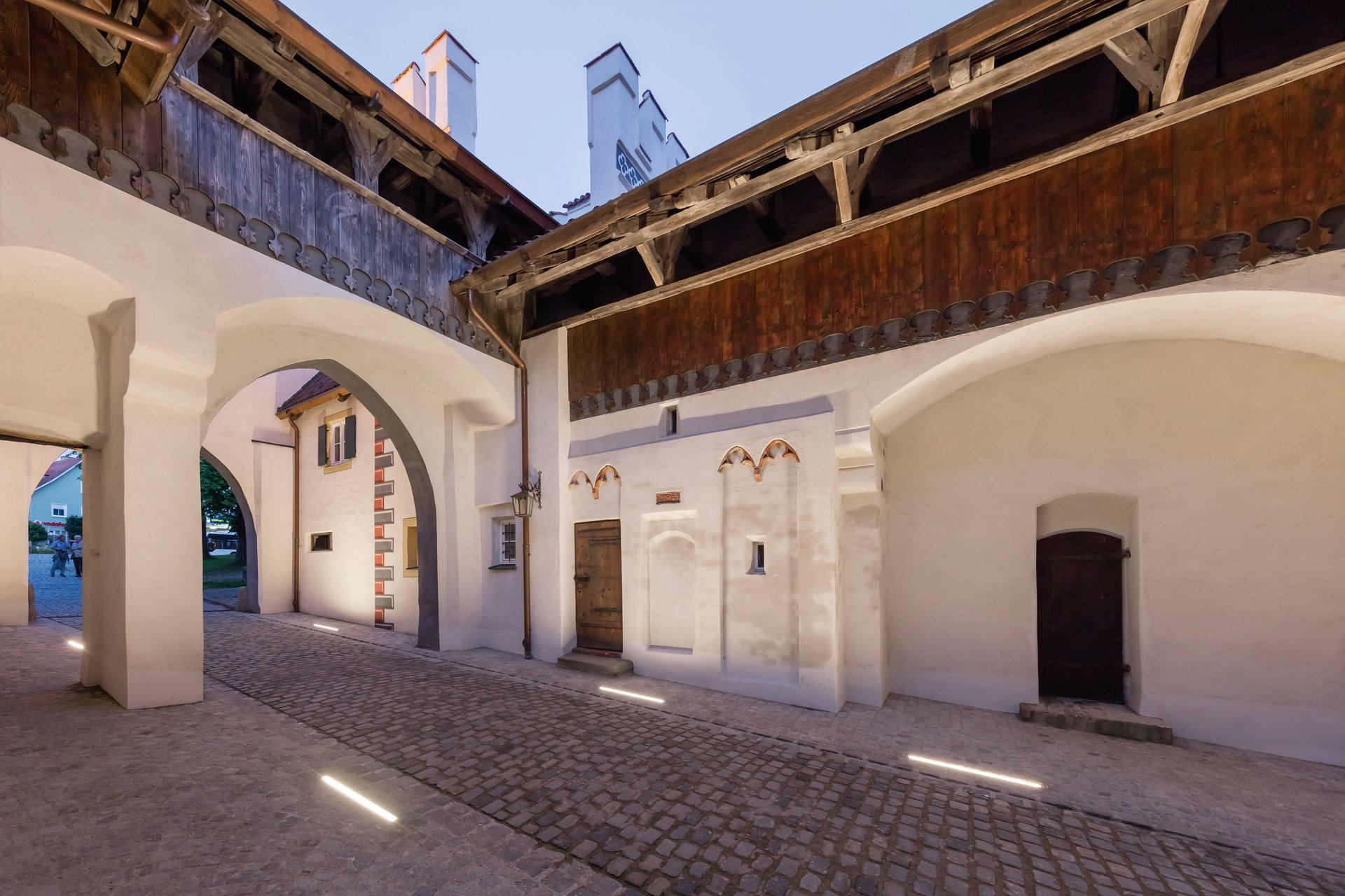 Bayertor fortress complex in Landsberg at blue hour showing complete Gothic barbican with white plastered walls, traditional timber-framed galleries, arched passageways, and WE-EF LED lighting system that enhances pedestrian safety while respectfully illuminating the 15th-century architecture