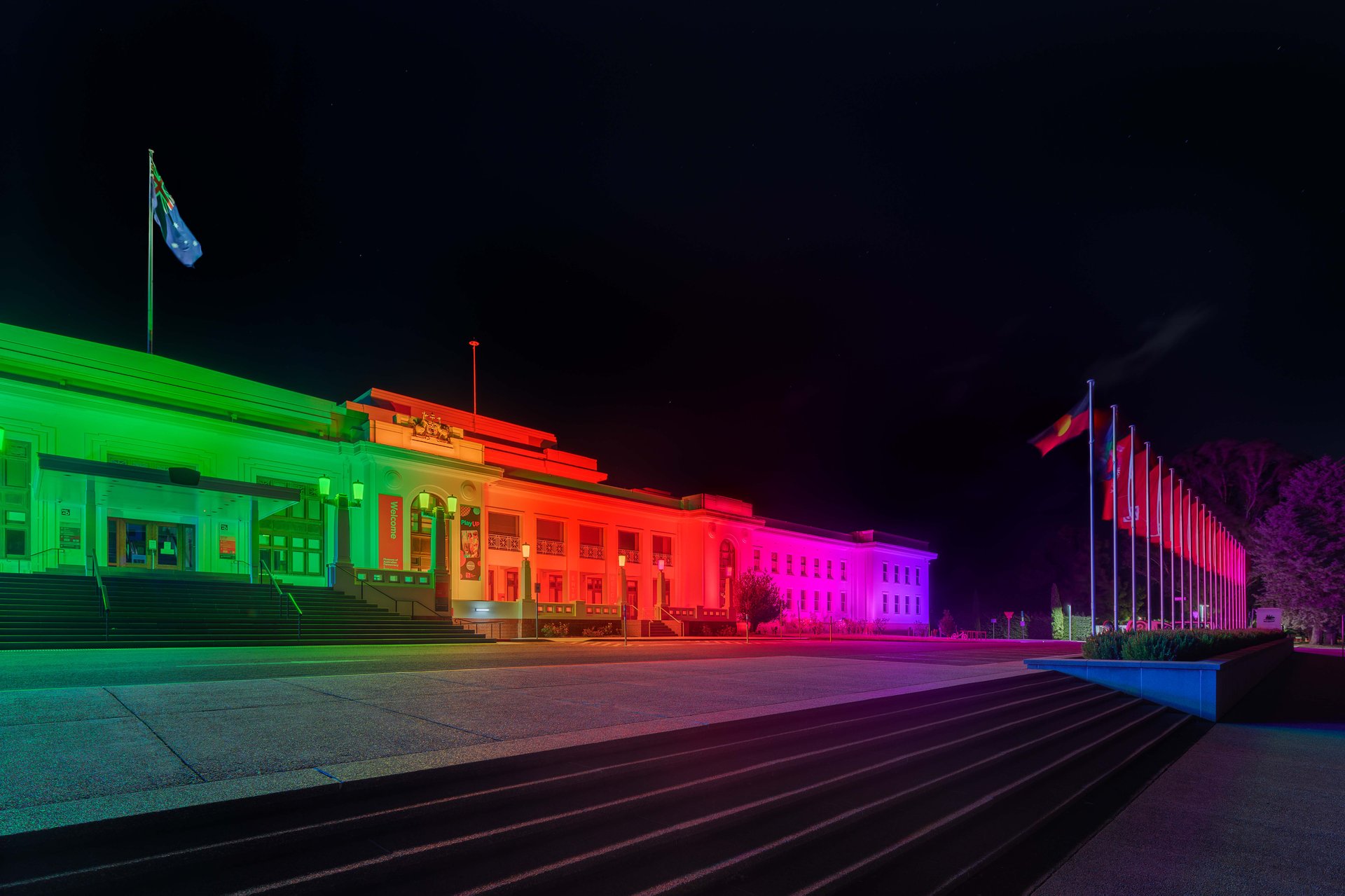Old Parliament House displaying complete rainbow spectrum from green through red, orange, yellow to magenta and purple demonstrating full RGBW and RGBA color-changing capability of WE-EF luminaires, dynamic lighting effects painting entire 1920s stripped classical government building in coordinated color wash, Australian flag illuminated on left, flagpoles visible on right, precise wallwash lens technology finessed by Integral Lighting team ensuring uniform coverage across architectural details, Museum of Australian Democracy Canberra