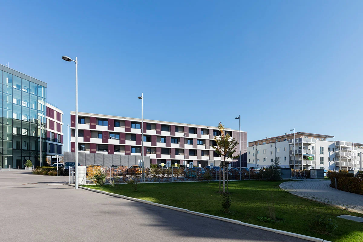 Modern residential and business development Q.West Ingolstadt showing contemporary mixed-use buildings with colorful checkerboard facade patterns, WE-EF street lighting poles, and landscaped pedestrian pathways under clear blue sky