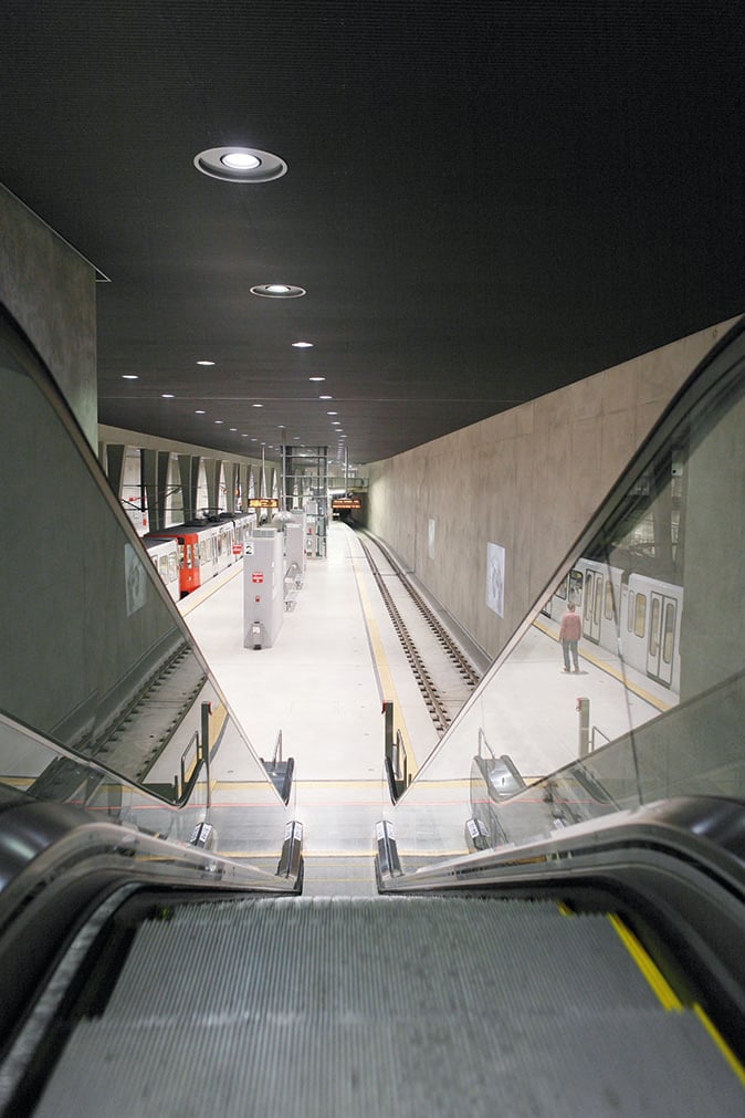 Top-down view of two escalators descending to the underground platform at Breslauer Platz station in Cologne, with WE-EF recessed ceiling luminaires and an incoming tram