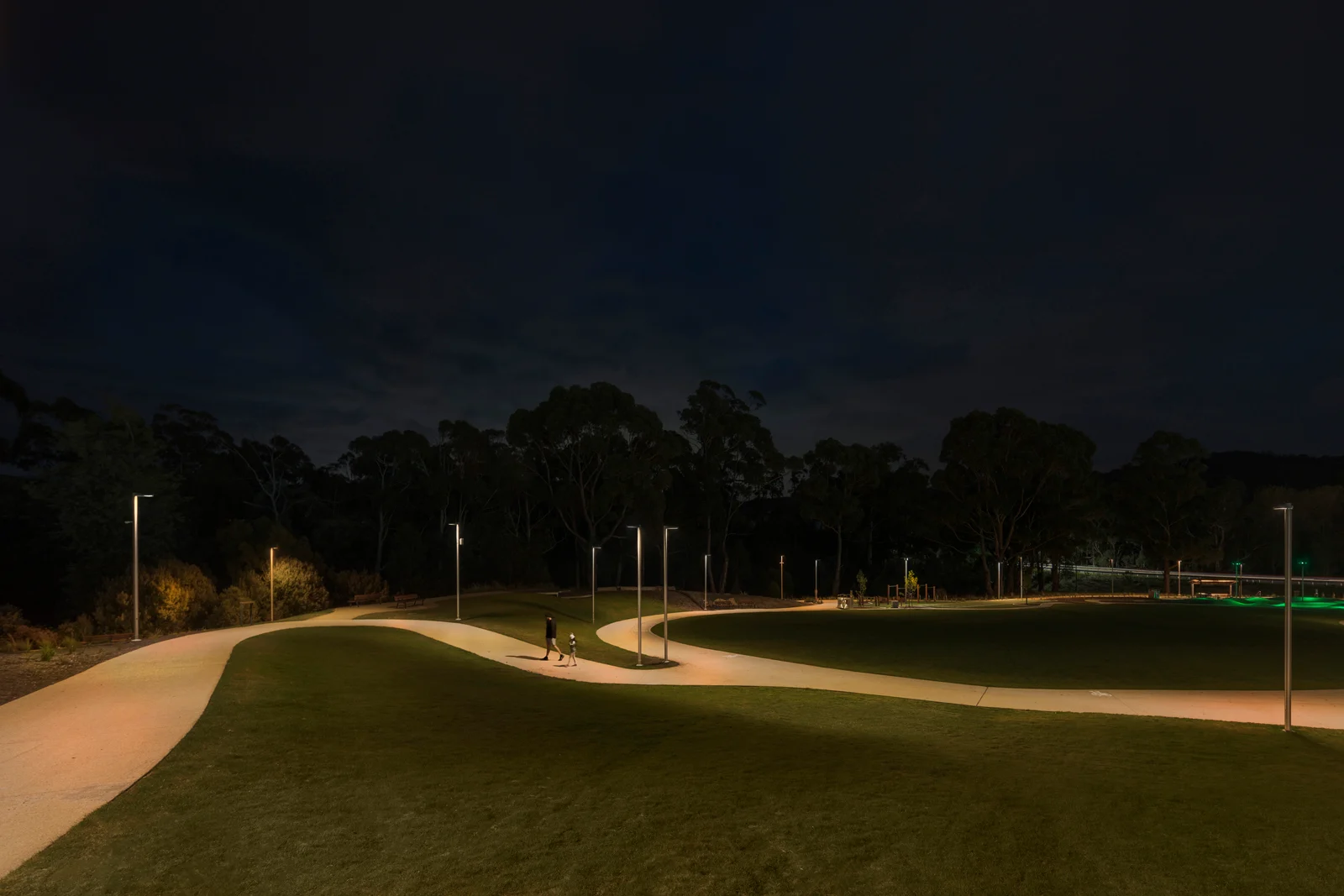 Kingston Parklands at night showing contrast between ultra-warm 2200K WE-EF VFL530 LED pathway lighting creating distinctive amber glow on Creek Pathway (left) and warmer 2700K playground area lighting (right), two visitors walking through landscape designed by Playstreet with native Tasmanian eucalyptus forest backdrop silhouetted against dark sky, demonstrating dual color temperature strategy and precise light distribution using P65 and S65 optics that minimizes light pollution while ensuring safe recreational use for Kingborough community near Hobart