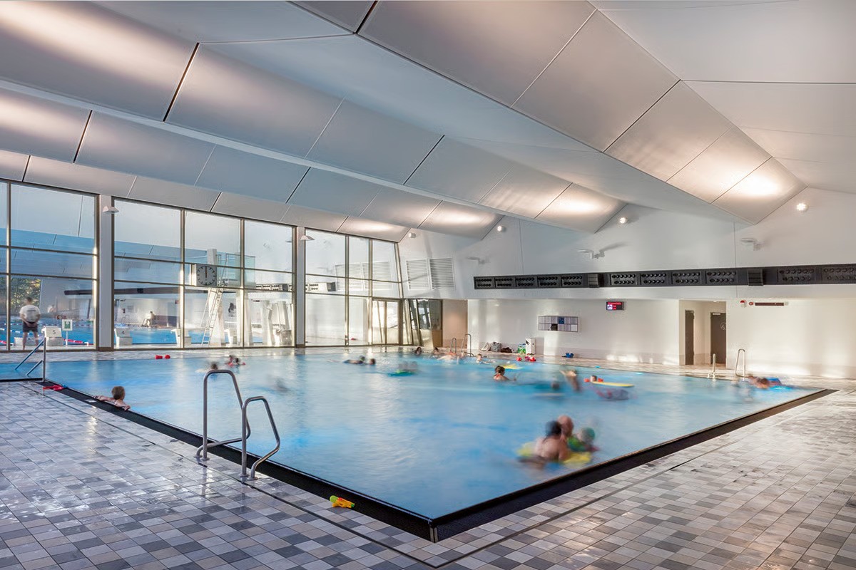 Hamburg-Ohlsdorf family swimming pool 50-meter dividable competition pool with WE-EF PIA240 compact floodlights with asymmetric forward-throw distribution mounted on pool partition providing homogenous illumination, PFL540 wall-bracket luminaires visible along hall sides, clear bright friendly atmosphere enhanced by comprehensive lighting concept using outdoor-rated luminaires with corrosion resistance for extreme swimming pool conditions, designed by IB Eneratio Hamburg technical building services planners