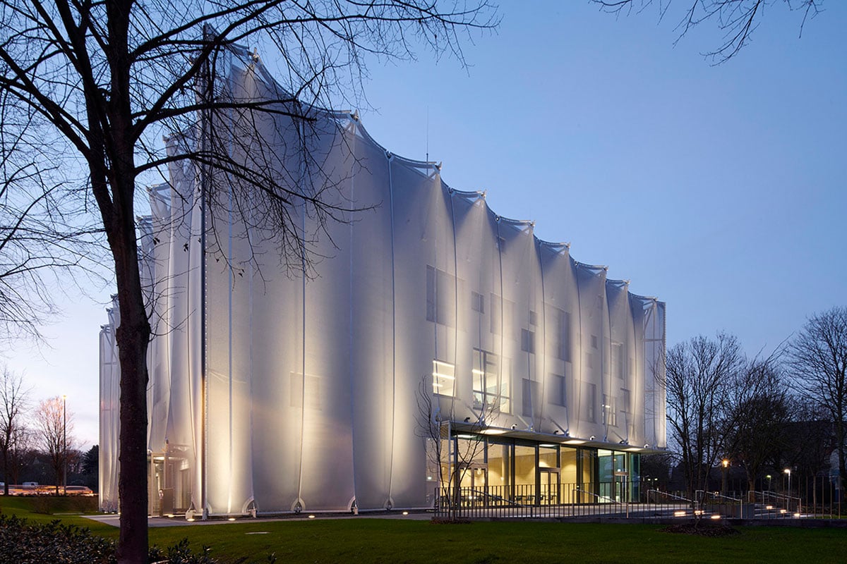 Textile Academy NRW at night showing comprehensive facade lighting by Maass-Licht Hamburg using WE-EF ETC140-GB gimbal-mounted inground luminaires and FLC141 projectors on entrance canopy, intensity-adjusted illumination allowing textile membrane transparency variation to showcase complex design language, large glass facade visible through sheath opening in entrance area welcoming students and visitors to private vocational college funded by regional textile industry associations in Mönchengladbach center of traditional German textile manufacturing