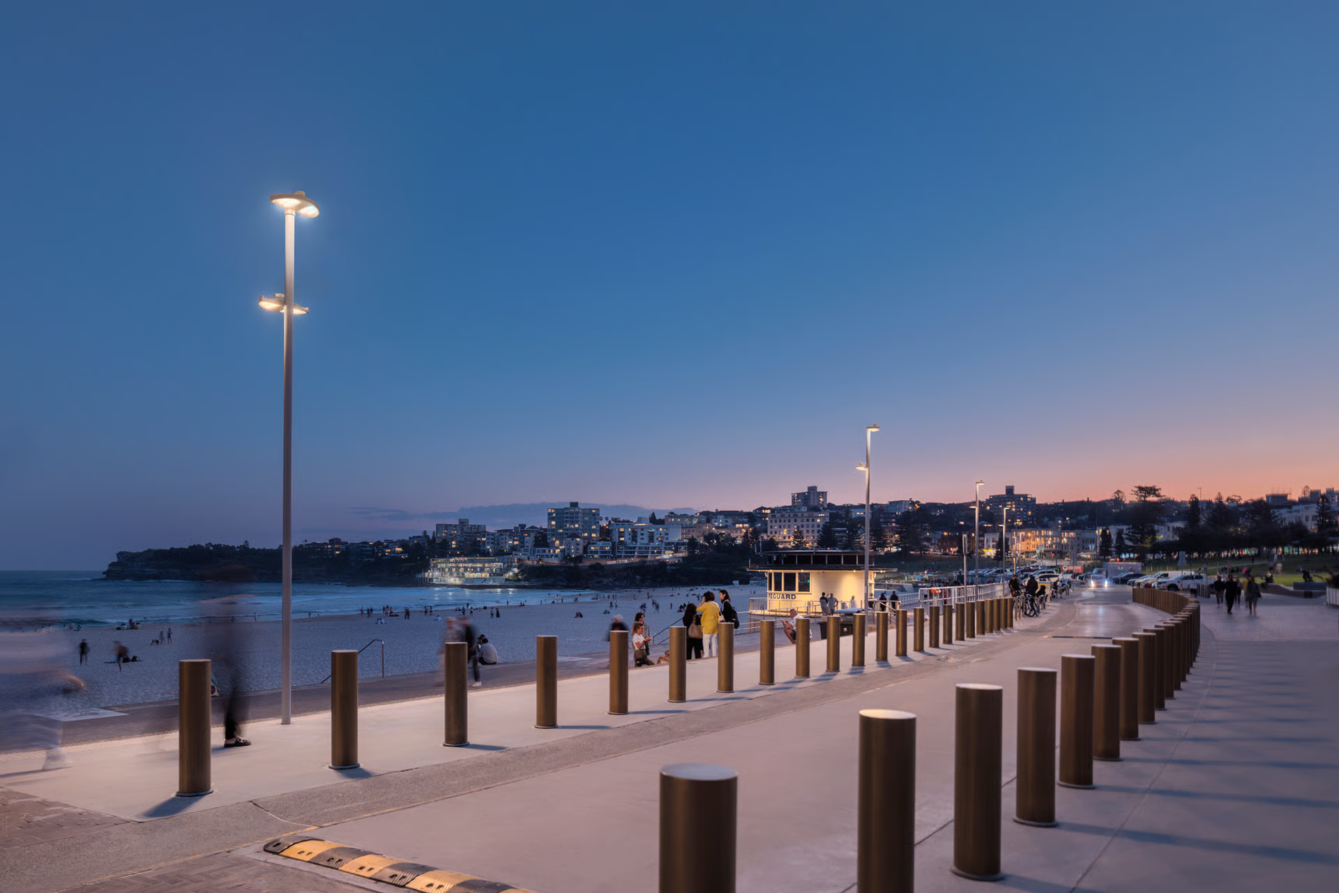Bondi Beach promenade at sunset showing reimagined parkland with WE-EF VLS400 pole-mounted LED luminaires providing warm 3000K lighting, rows of bronze bollards delineating pedestrian and cycling zones, iconic yellow lifeguard tower, Bondi headland and residential buildings in background, demonstrating 5-star sustainable design and salt-resistant luminaires chosen by Waverley Council for 25+ year longevity in harsh coastal environment