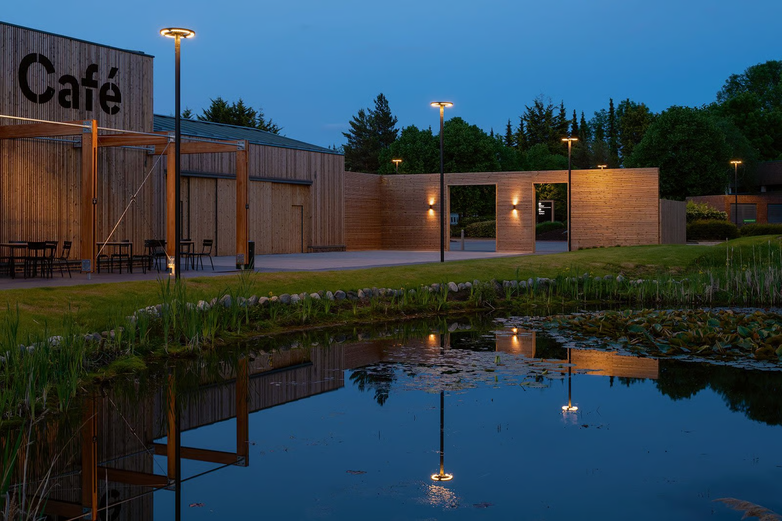Croxley Park office campus The Hive service centre at twilight near Watford showing pond reflection and WE-EF CFT540 ring-shaped LED luminaire on 5-meter aluminum pole with warm 3000K illumination, three-year transformation by Columbia Threadneedle of 30+ hectare parkland site north of London integrating workplace with living space through café, gym, day-care amenities and comprehensive outdoor lighting by ESA architects creating pleasant feel-good atmosphere enabling employees to comfortably work late and enjoy campus beyond office hours