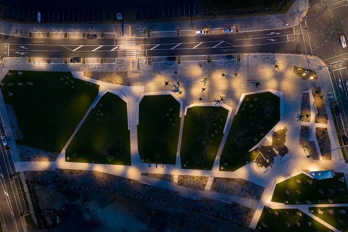 Aerial night view of Te Hauāuru Park Westgate showing organic lawn patterns structured by walls inspired by upper Waitematā harbour inlets, innovative WE-EF lighting design by MHL Auckland using 66 FLC121 projectors with medium-beam optics mounted at varying heights (5.5-6.5m) on 7-meter poles creating directional pinpointed illumination avoiding standard grid layout, P2 standard lighting ensuring safe thoroughfare between North West Shopping Centre (one of Australasia's largest precincts) and apartments in rapidly developing Auckland town centre 20km west of city