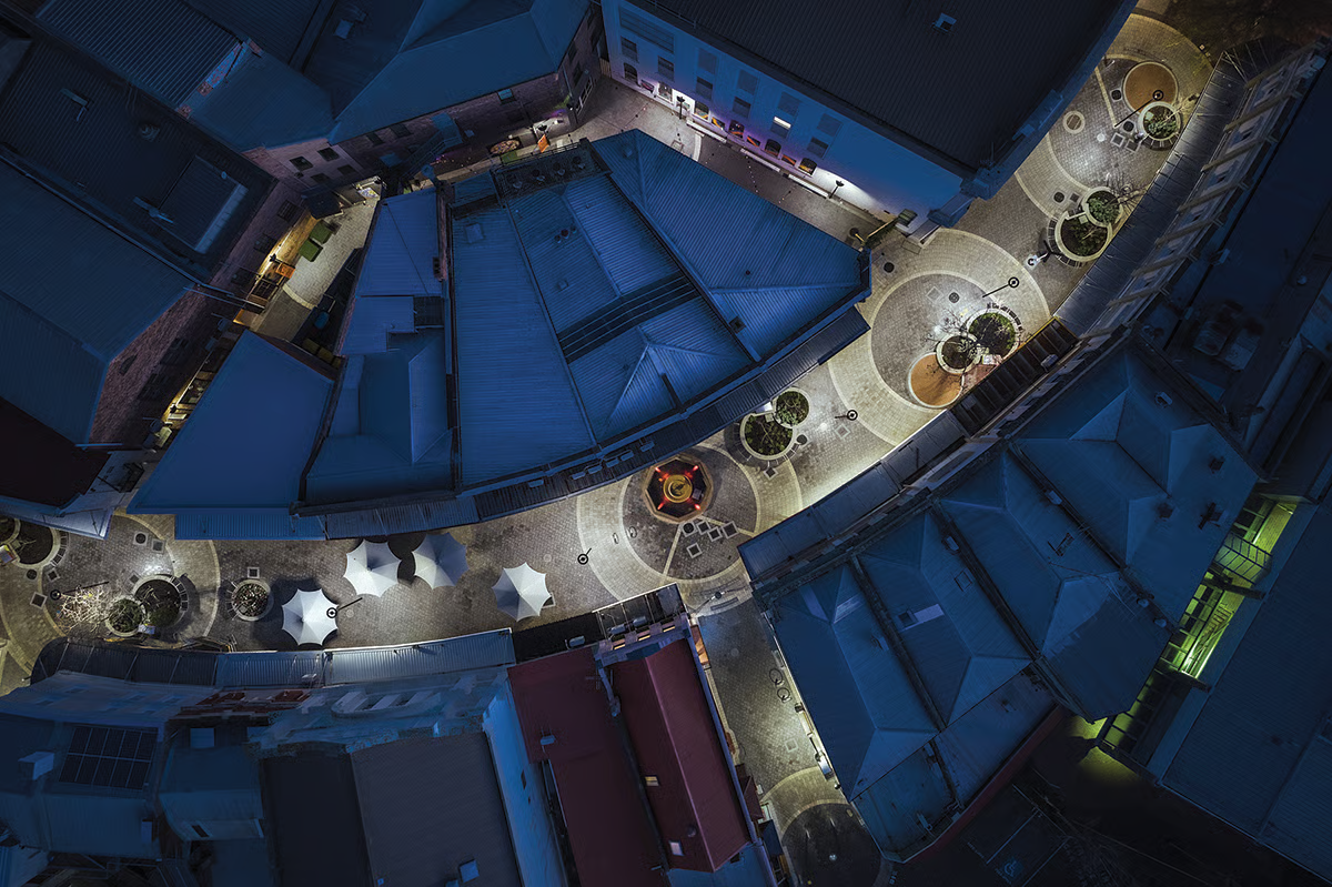 Aerial view of Quadrant Mall at night showing complete urban renewal with WE-EF CFT540 LED lighting strategically positioned along pedestrian zones, circular planters, event stages with white canopy umbrellas, fountains, and graphic paving patterns creating an inviting retail and entertainment hub in Launceston's city center