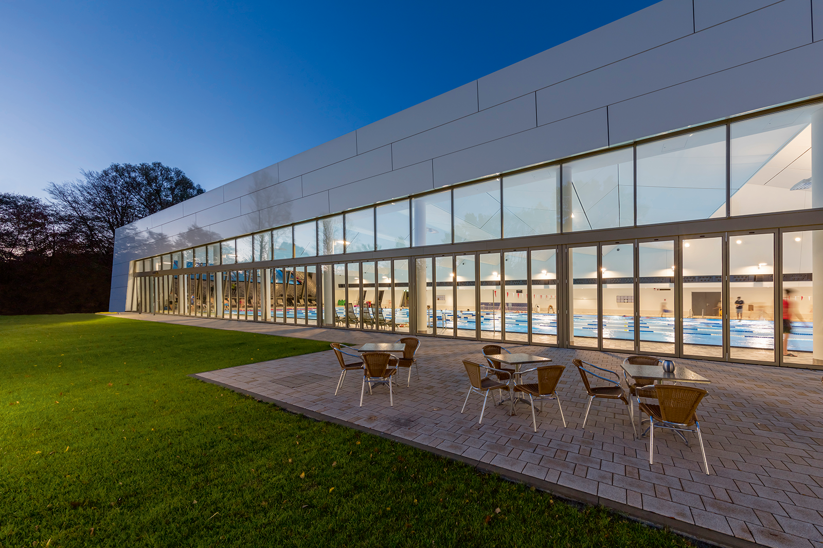 Hamburg-Ohlsdorf family swimming pool exterior at dusk showing prismatic shimmering metallic structure with simple futuristic design by Czerner Göttsch Architects, long glazed south facade that fully opens in fine weather connecting indoor all-weather sports pool with outdoor sunbathing lawn, comprehensive WE-EF LED lighting system including PFL540 wall-bracket luminaires, PIA240 compact floodlights, and FLC230 ceiling projectors creating sustainable attractive durable facility with warm 3000K lighting throughout using 5CE Superior Corrosion Protection technology for chlorinated pool environment