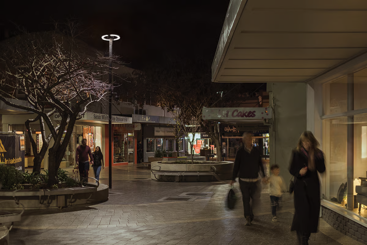 ImageQuadrant Mall at night with families and shoppers enjoying the pedestrian zone, illuminated by WE-EF CFT540 LED pole lights with integrated motion sensors and OLC lens technology providing glare-free lighting, circular planters with winter trees, and active storefronts including optometrist and cake shop