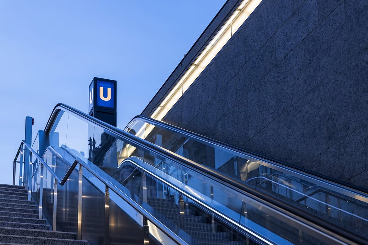 Image (Station entrance detail with escalators): "Museumsinsel U-Bahn station entrance at blue hour showing contemporary design without traditional Berlin underground light portal, open granite parapet with WE-EF VLR100 linear wall luminaires integrated into transparent covered wall boxes providing asymmetric wall wash downlighting, glass-enclosed escalators leading to 180-meter platform beneath Spree Canal, iconic blue U-Bahn signage, designed by Max Dudler Architects as underground entrée to Museum Island cultural institutions completing U5 line connection between Brandenburger Tor and Alexanderplatz